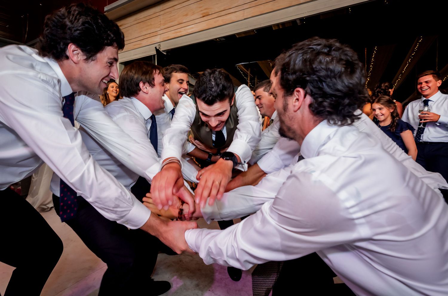 Groomsmen in white shirts huddle together laughing and celebrating on a wedding dance floor.