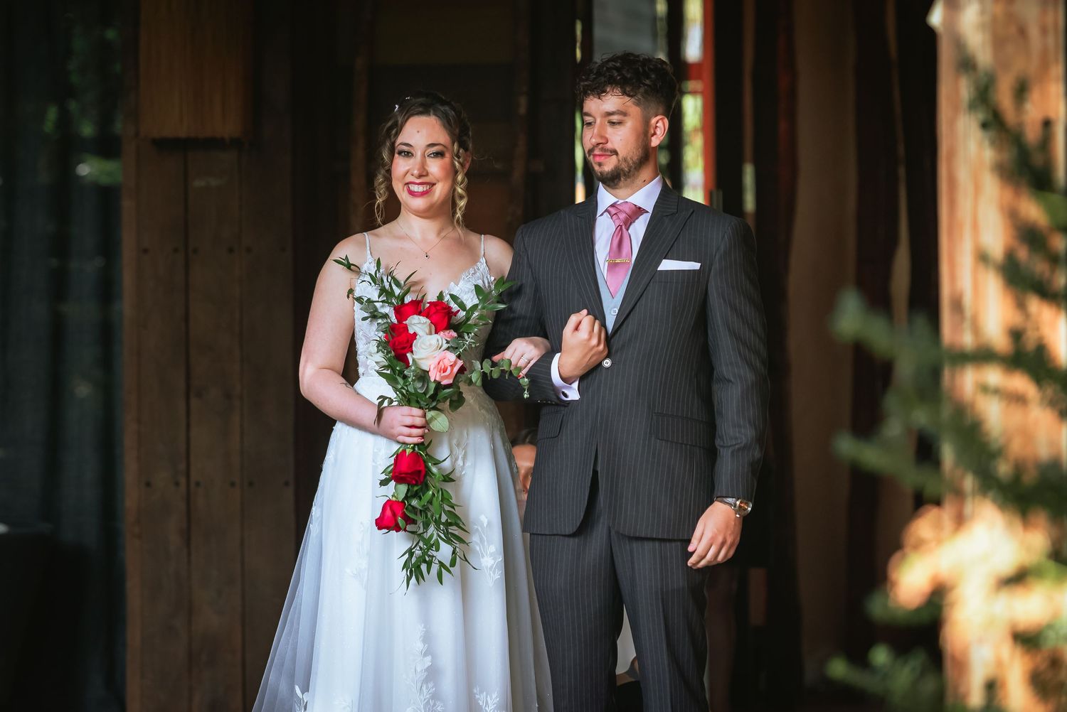 Bride in white gown holding red bouquet stands with groom in gray suit at wooden venue entrance.