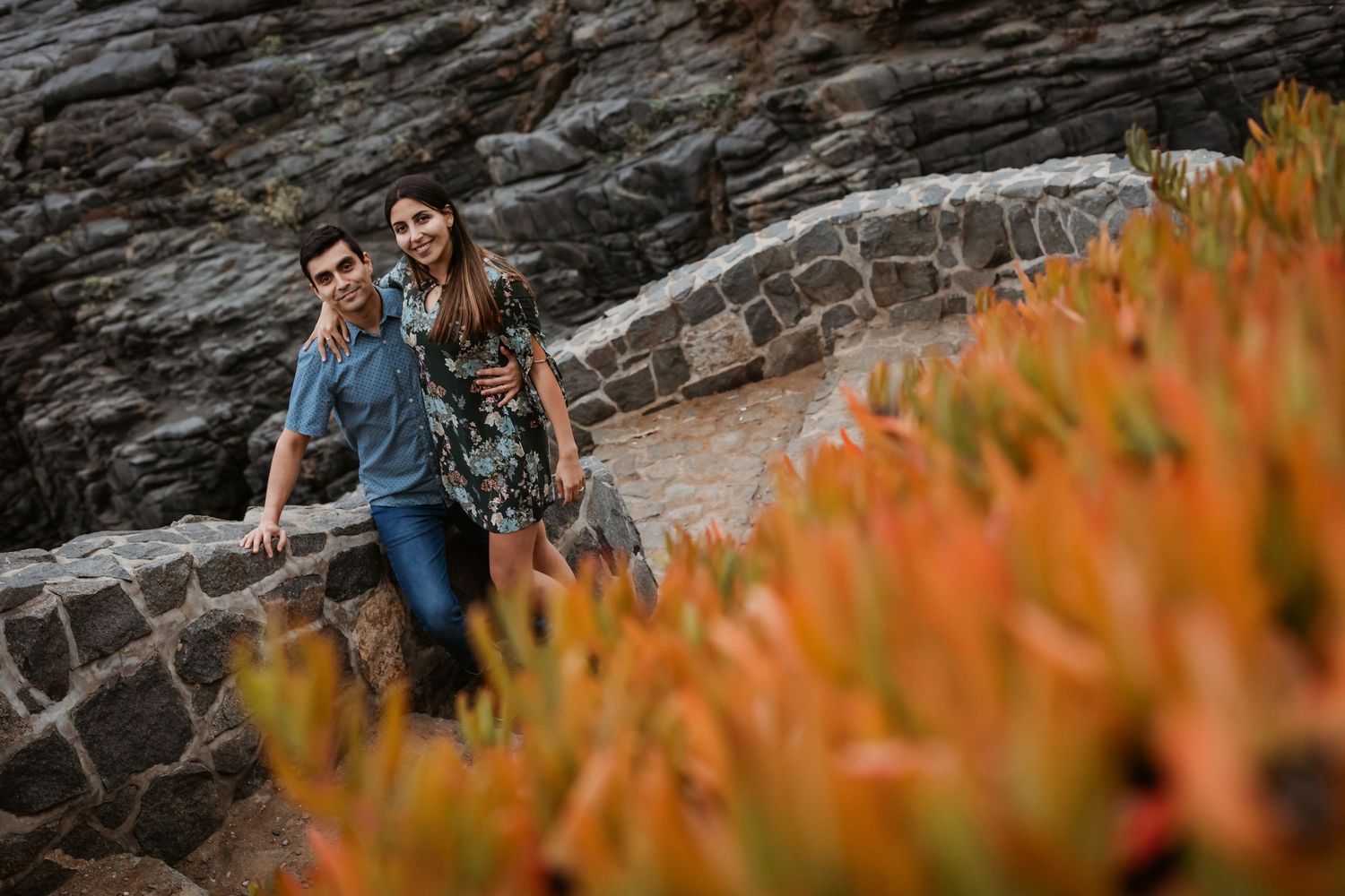 Smiling couple leaning on stone wall surrounded by colorful coastal succulents.