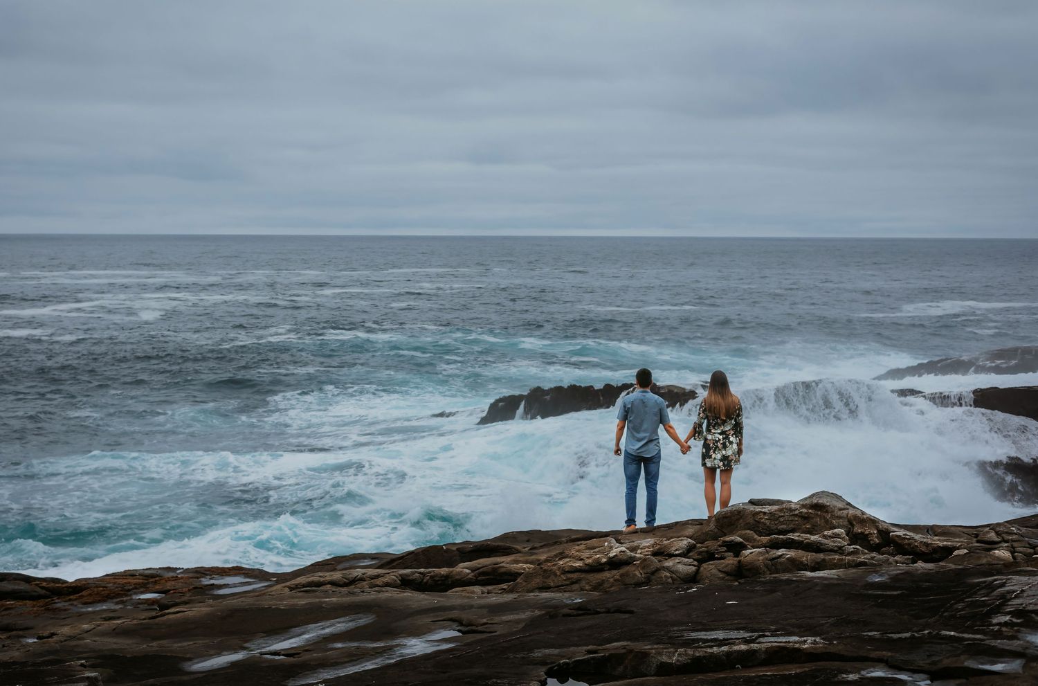 Couple standing hand in hand viewing powerful ocean waves from rocky coastal overlook.