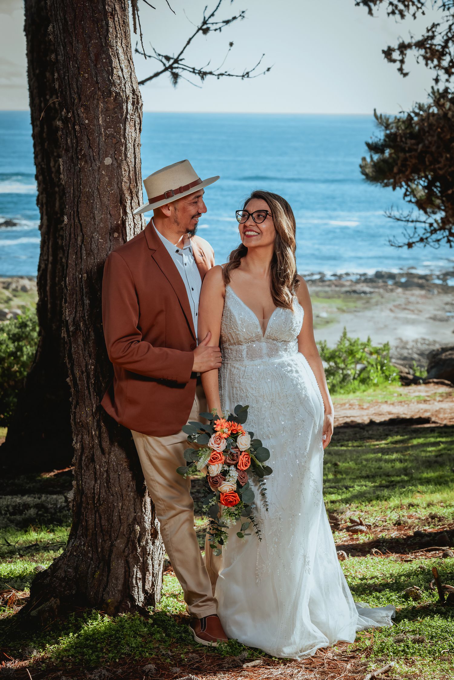 Smiling bride in a white lace gown holding a bouquet leans against a tree beside her groom in a straw hat, with the ocean behind them.