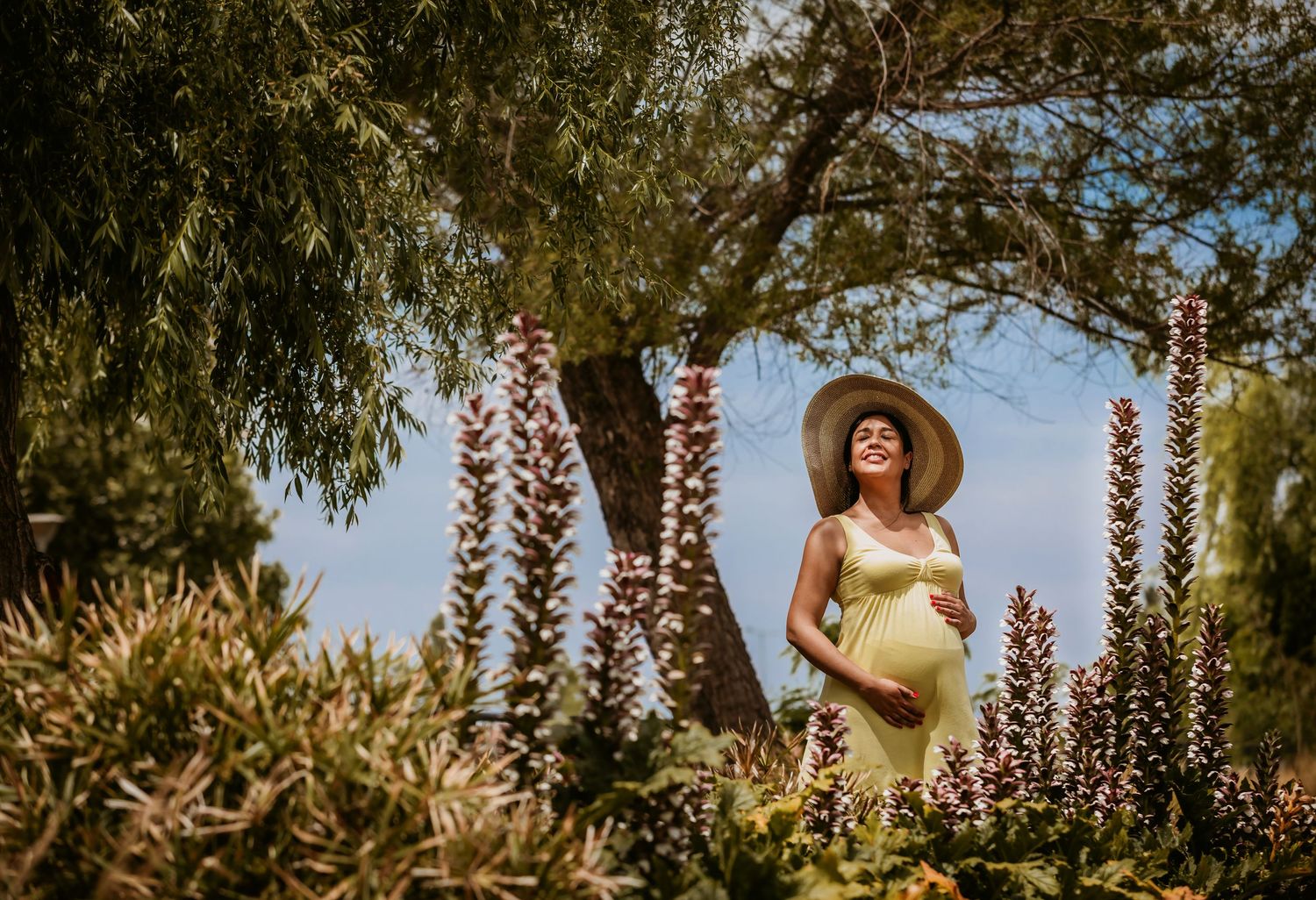 Smiling pregnant woman in yellow dress and sun hat standing among tall flowering plants in a sunny garden.