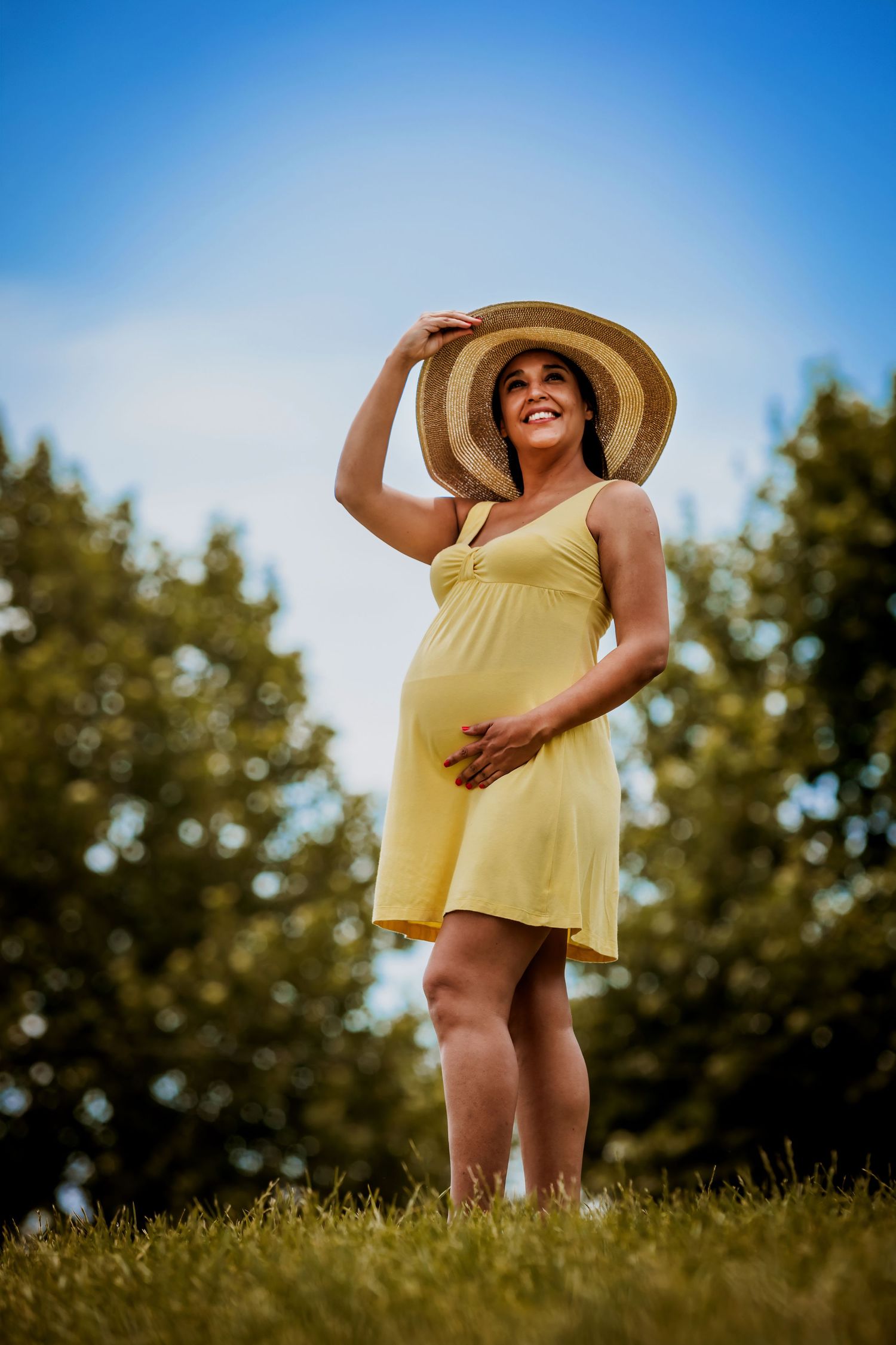 Smiling pregnant woman in yellow dress and straw hat standing on grass with trees in background.