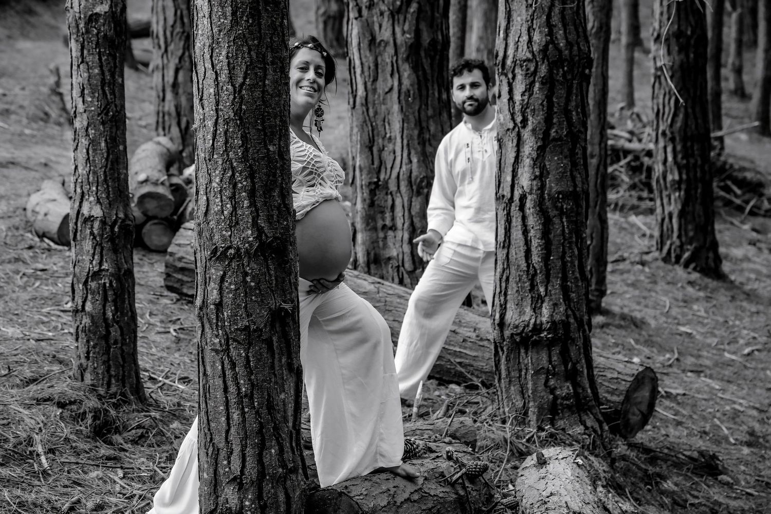 Black and white maternity photo of a pregnant couple standing among tall trees in a forest, wearing white outfits.