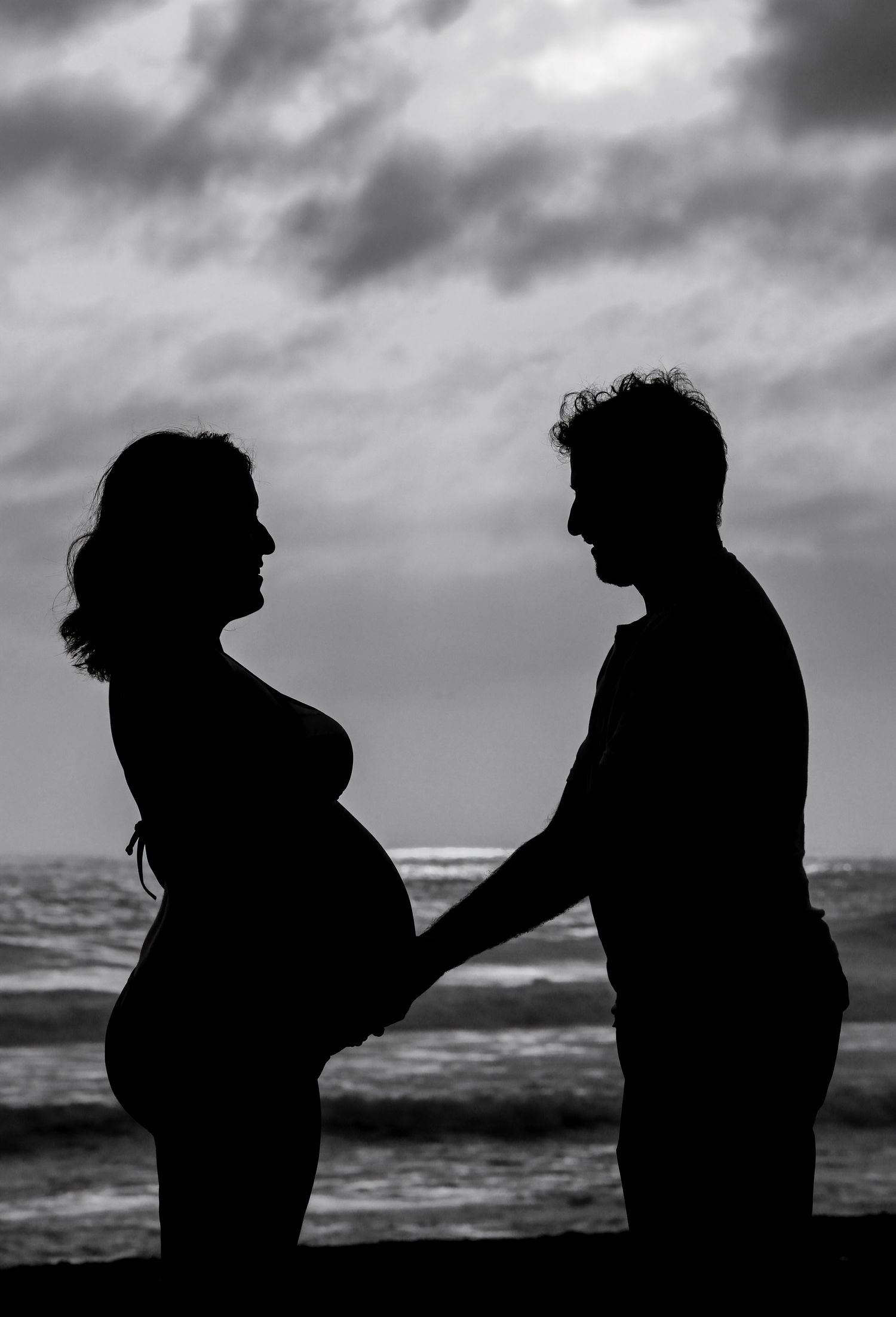 Black and white silhouette of a pregnant couple holding hands on a beach at sunset.