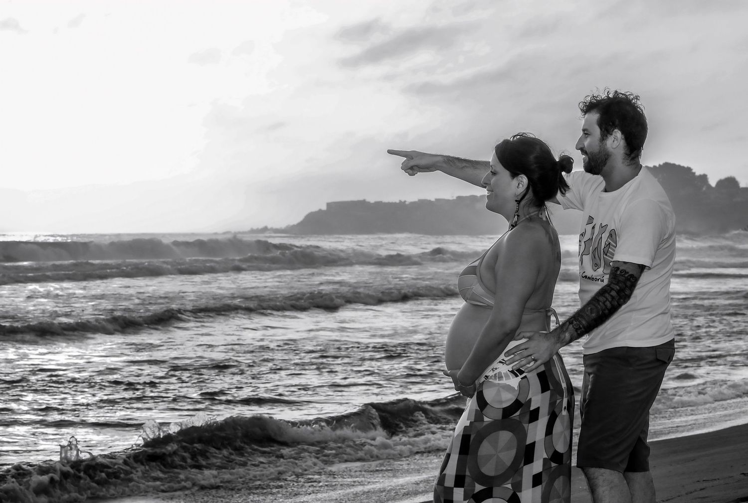 Pregnant couple standing on a beach in black and white, man pointing at ocean waves with rocky cliffs in background.