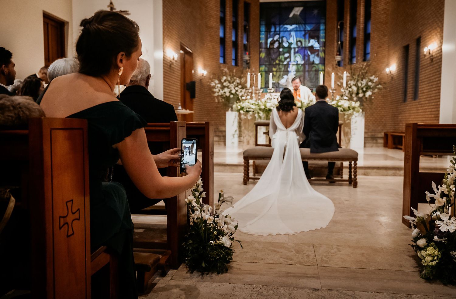A bride and groom kneel at the altar during a candlelit church wedding ceremony while a guest photographs the moment.