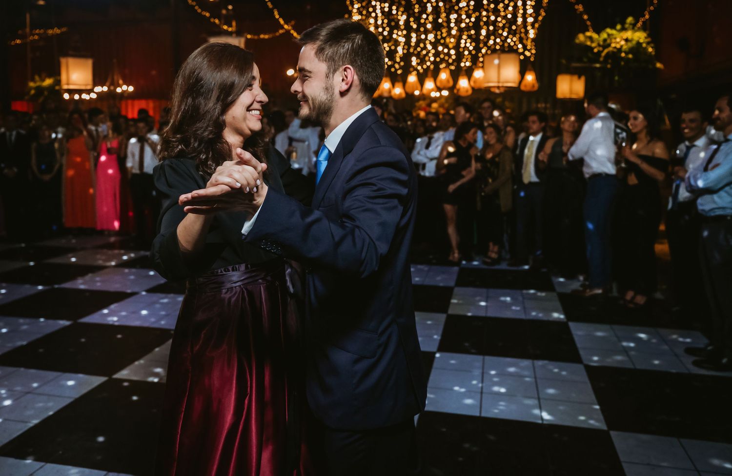 Couple sharing a romantic dance on a checkered floor at an elegant wedding reception with warm string lights.