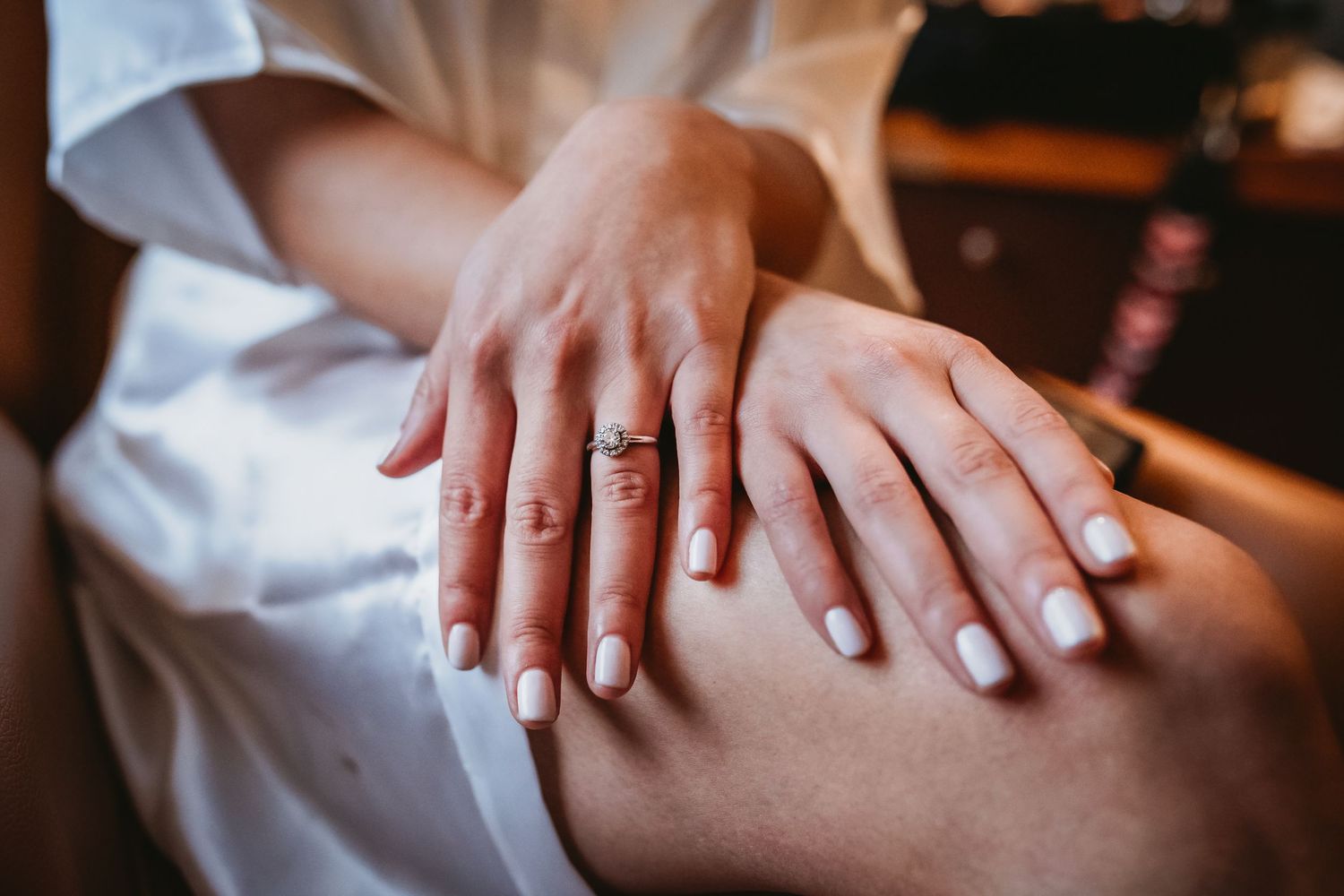 Close-up of a woman's hands with white manicure and elegant engagement ring resting on her knee.
