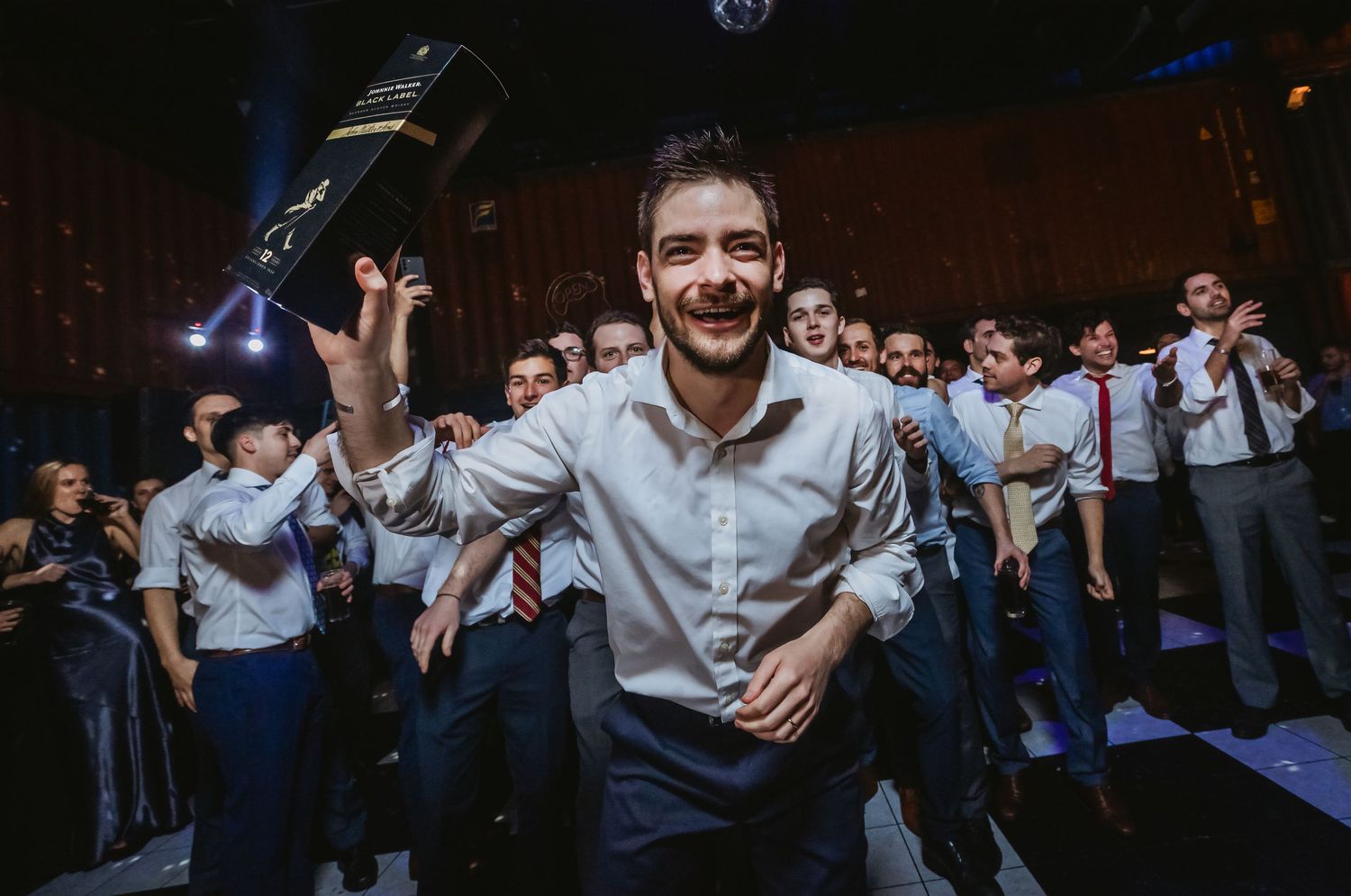 Young man celebrating at a party, holding a bottle aloft and smiling, surrounded by guests on a dance floor.