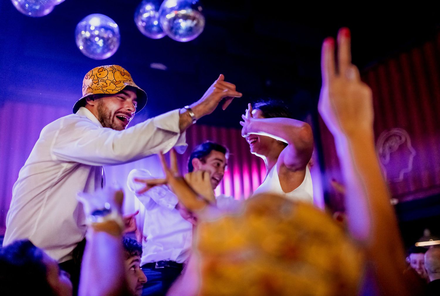 Man in yellow hat dancing energetically at a lively nightclub party with disco balls overhead.