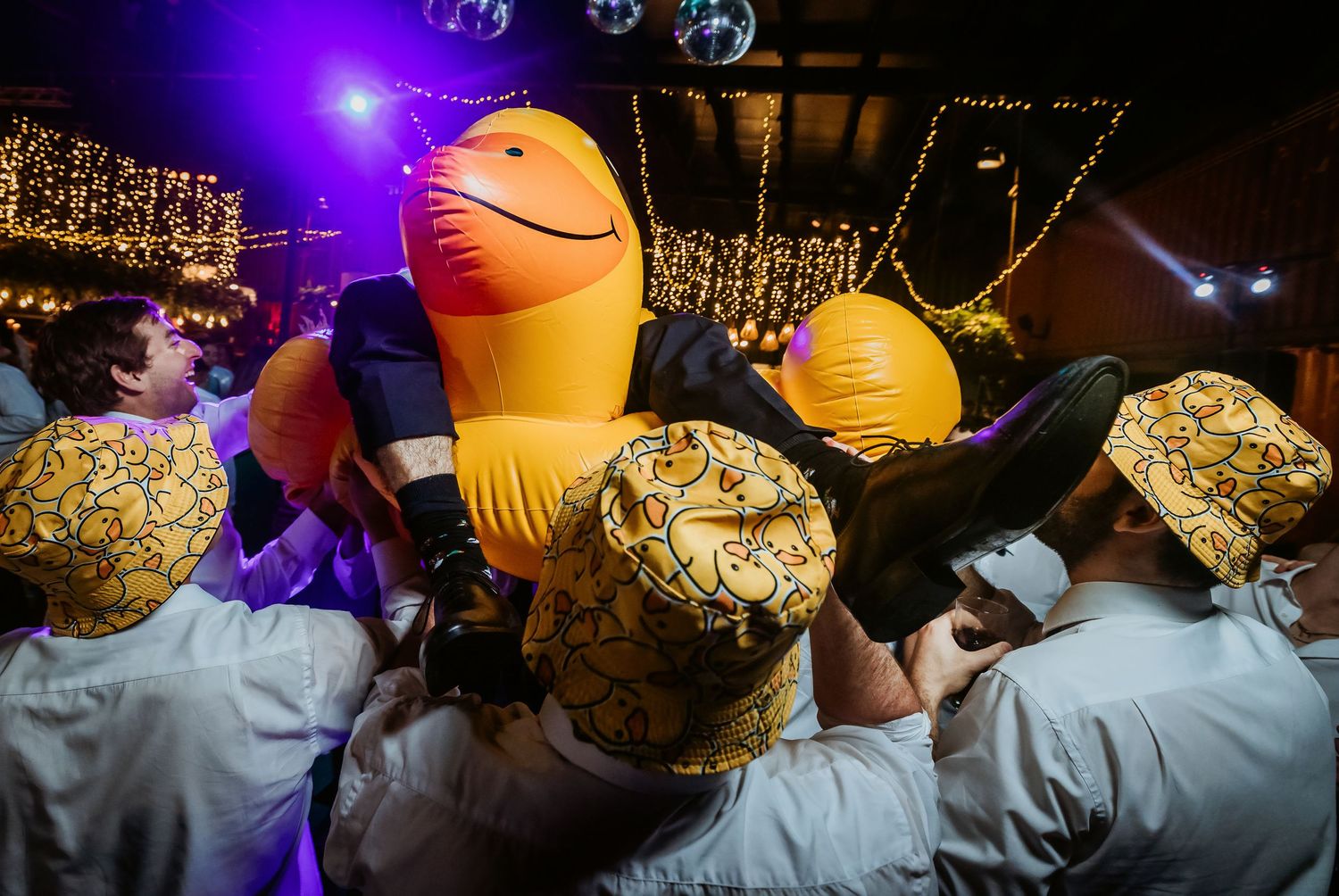 Party crowd in matching hats cheering around a giant inflatable toy under warm string lights at a lively indoor event.