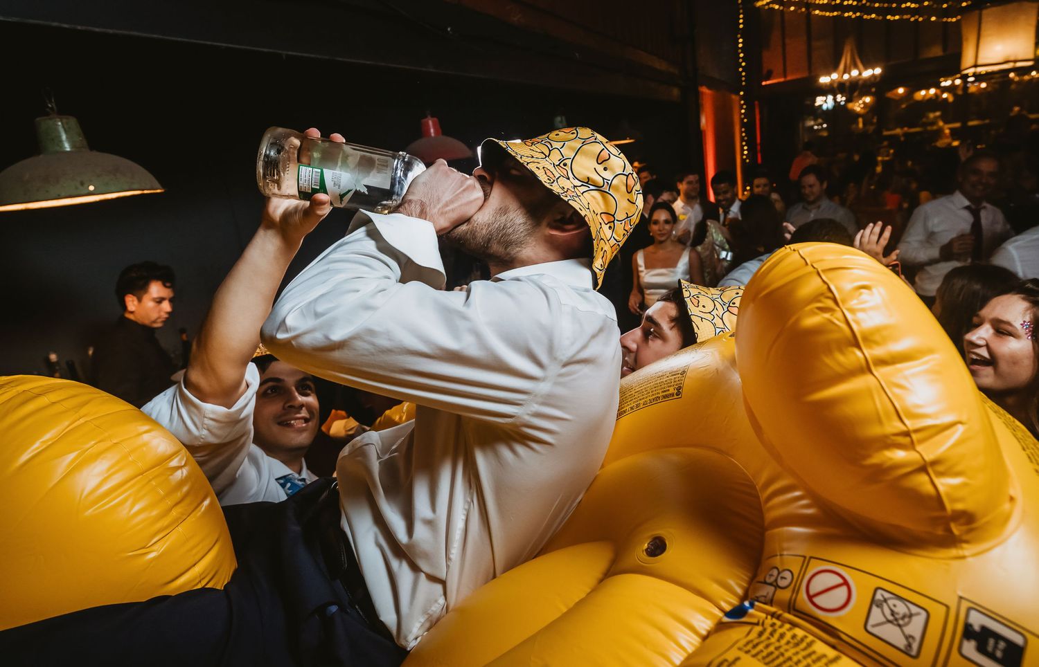 Man in bucket hat drinking from bottle beside inflatable yellow duck at crowded party.