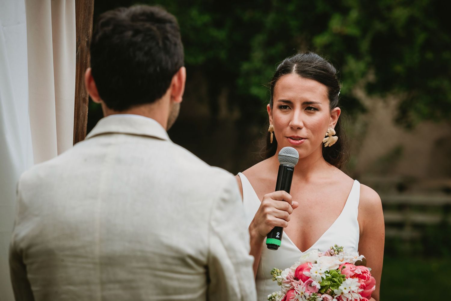 Bride reciting wedding vows into microphone wearing white dress with floral bouquet at outdoor ceremony.