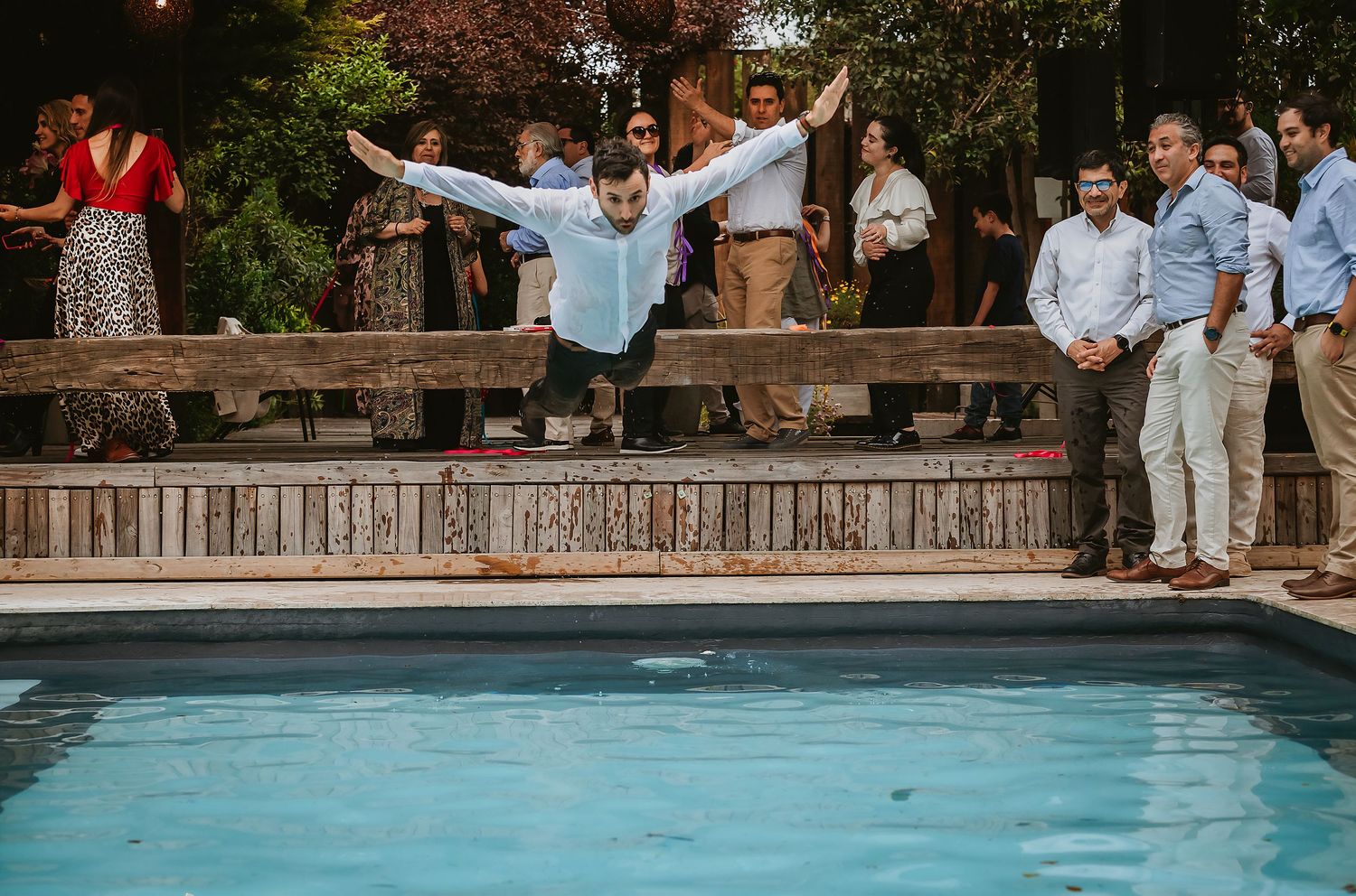 Man in blue shirt diving into pool at outdoor party with guests watching near wooden deck.