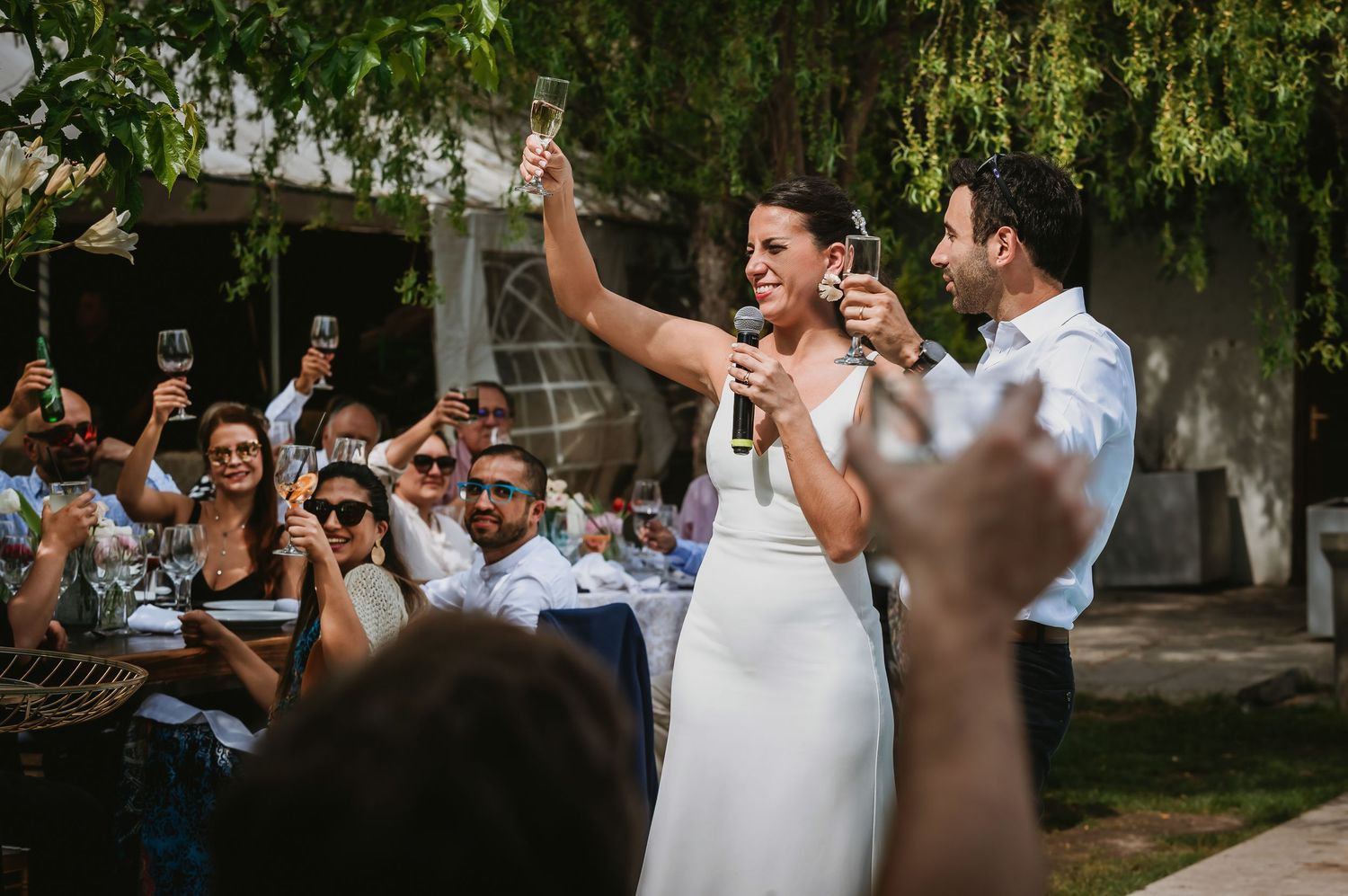 Bride and groom toasting with champagne glasses at outdoor wedding reception surrounded by cheering guests.