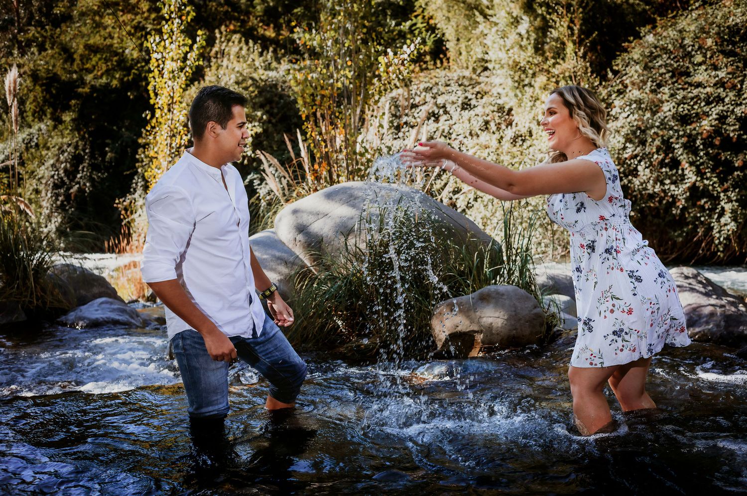 Couple playing in a rocky stream, woman splashing water at man, both smiling outdoors in nature.