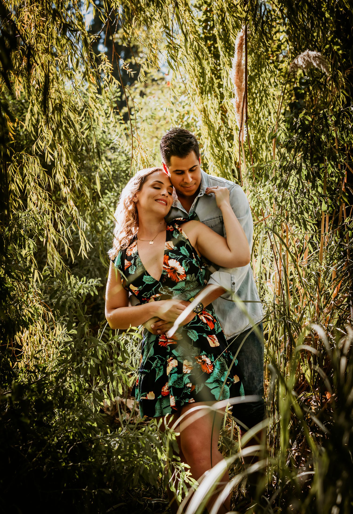 Couple embracing romantically among lush green weeping willow trees, woman in floral dress smiling softly.