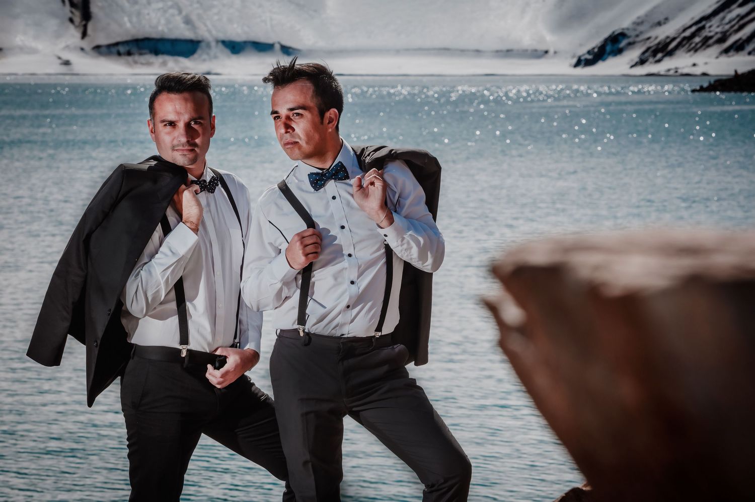 Two grooms in white shirts and suspenders pose confidently by a glacial lake and snowy peaks.