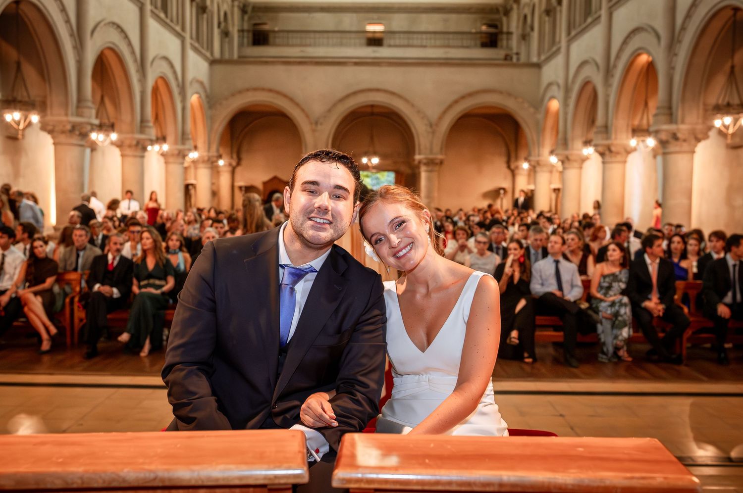 Smiling bride and groom pose together at church pew with wedding guests filling the background.