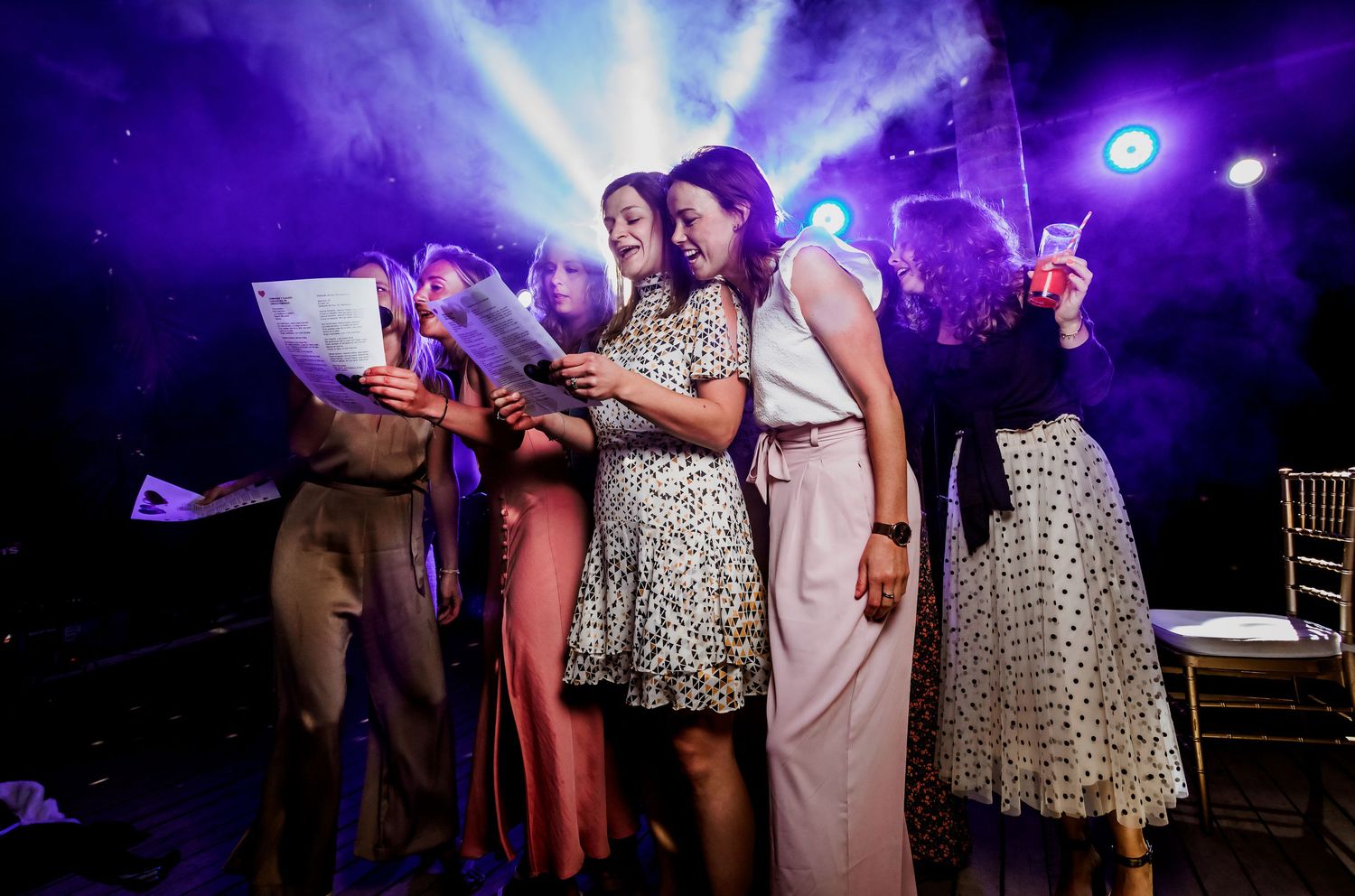 Bridesmaids singing together on stage under purple spotlights at wedding reception entertainment.