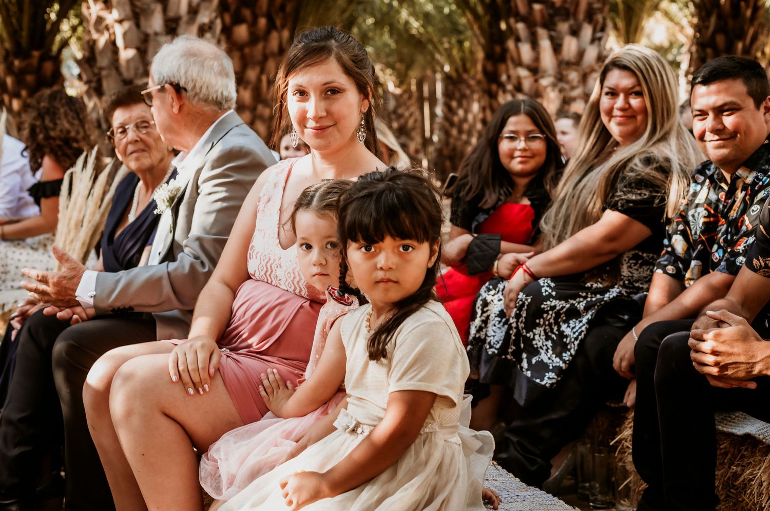 Family with young children seated together watching outdoor wedding ceremony under palm trees.