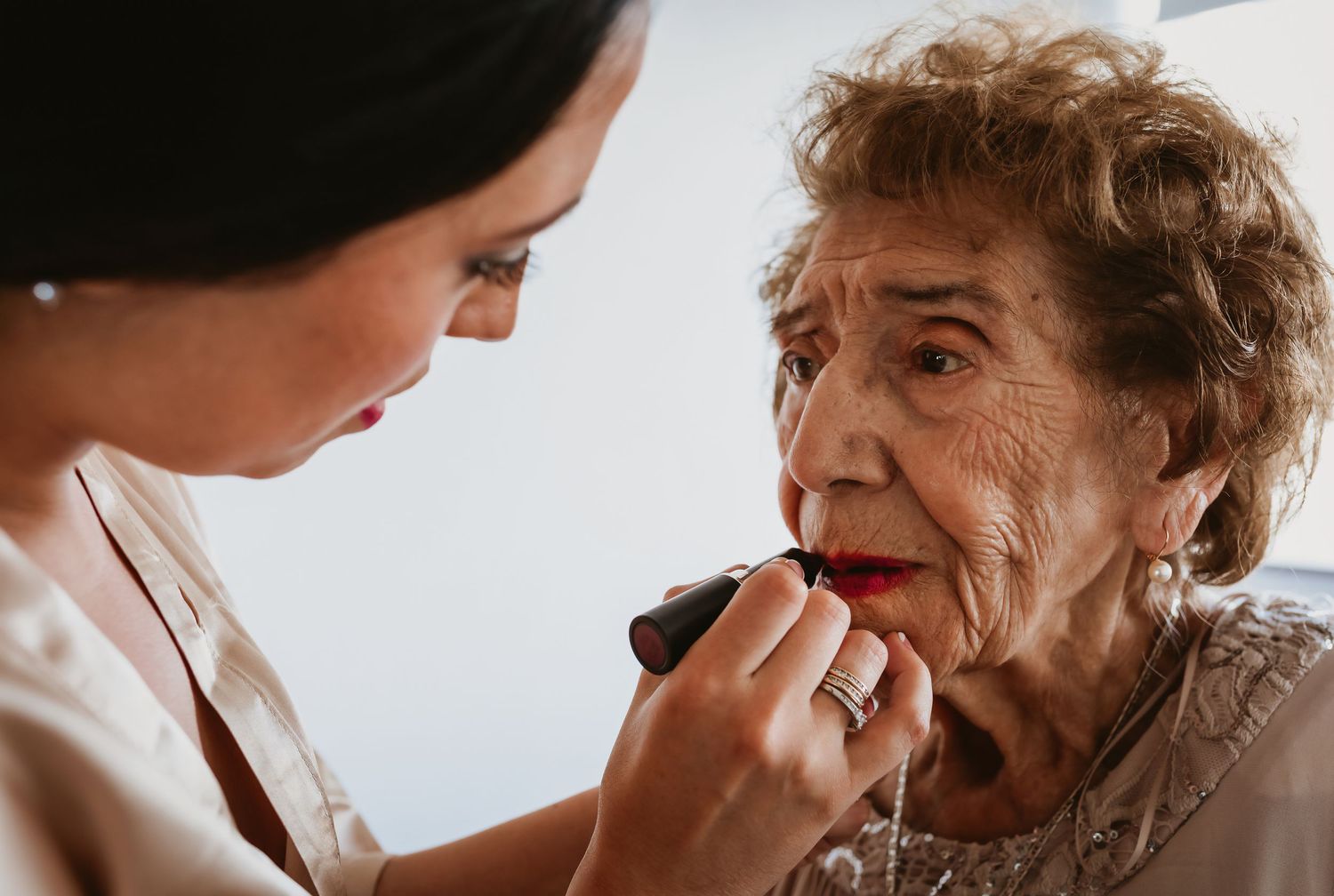 Young woman applying red lipstick to an elderly woman during bridal wedding preparations.
