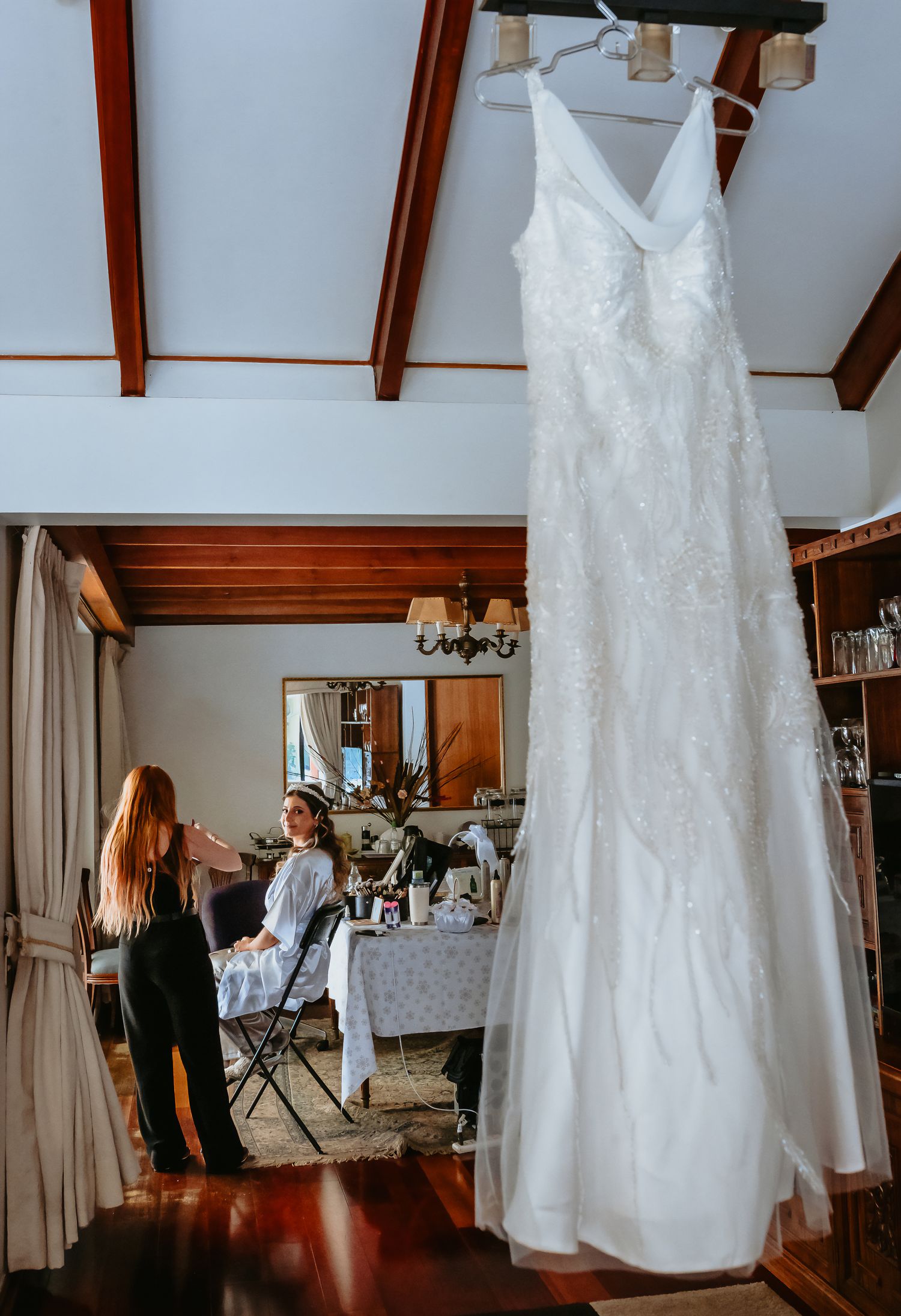 White lace wedding dress hanging in a rustic room while a bride gets ready in the background.