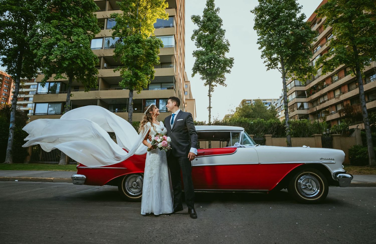 Bride and groom posing beside a red and white vintage classic car with bridal veil flowing in the wind.