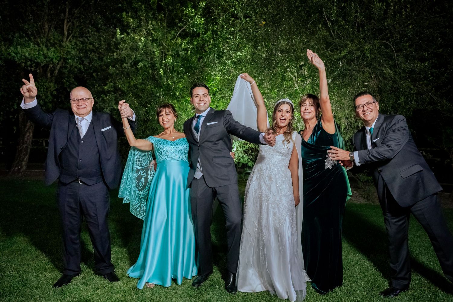 Joyful wedding party group photo with bride, groom, and family celebrating outdoors at night.