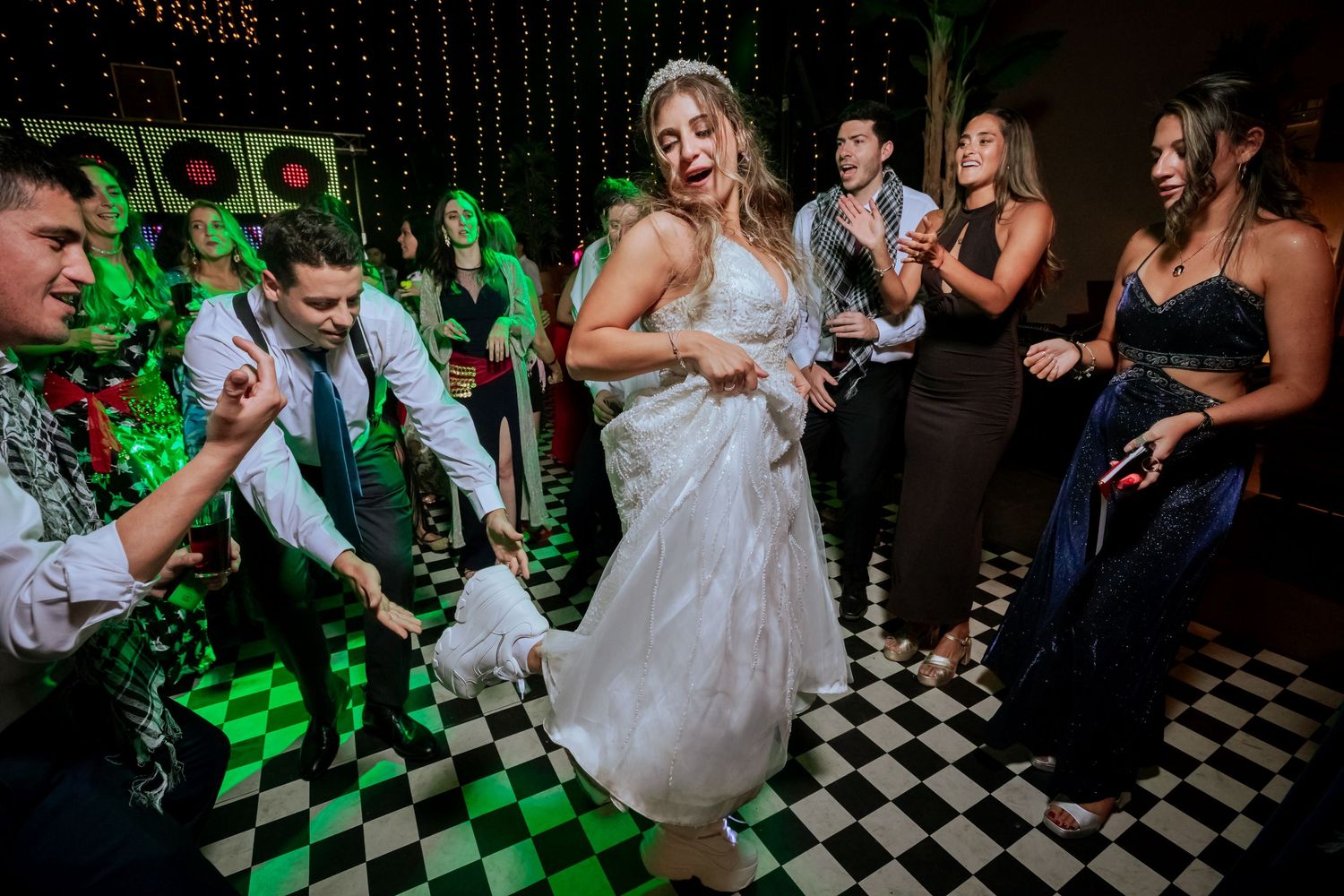 Bride dances enthusiastically in her wedding gown surrounded by celebrating guests on a checkered dance floor.