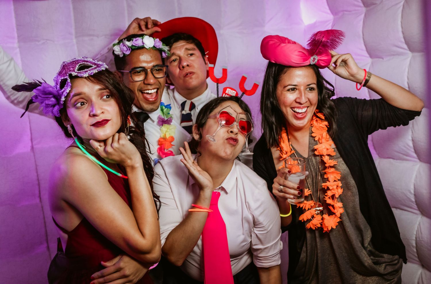 Five wedding guests pose with fun props and accessories in a glowing white photo booth at a reception.