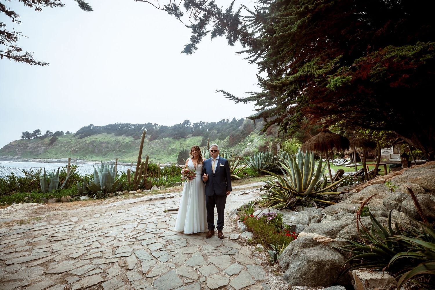 Bride and groom walk along a stone path with ocean views and cypress trees.