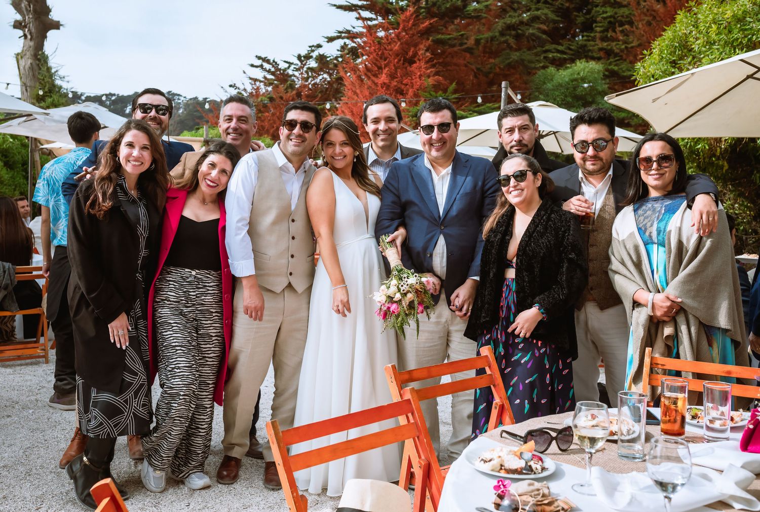 Wedding party group photo outdoors, bride in white dress with bouquet surrounded by smiling guests.