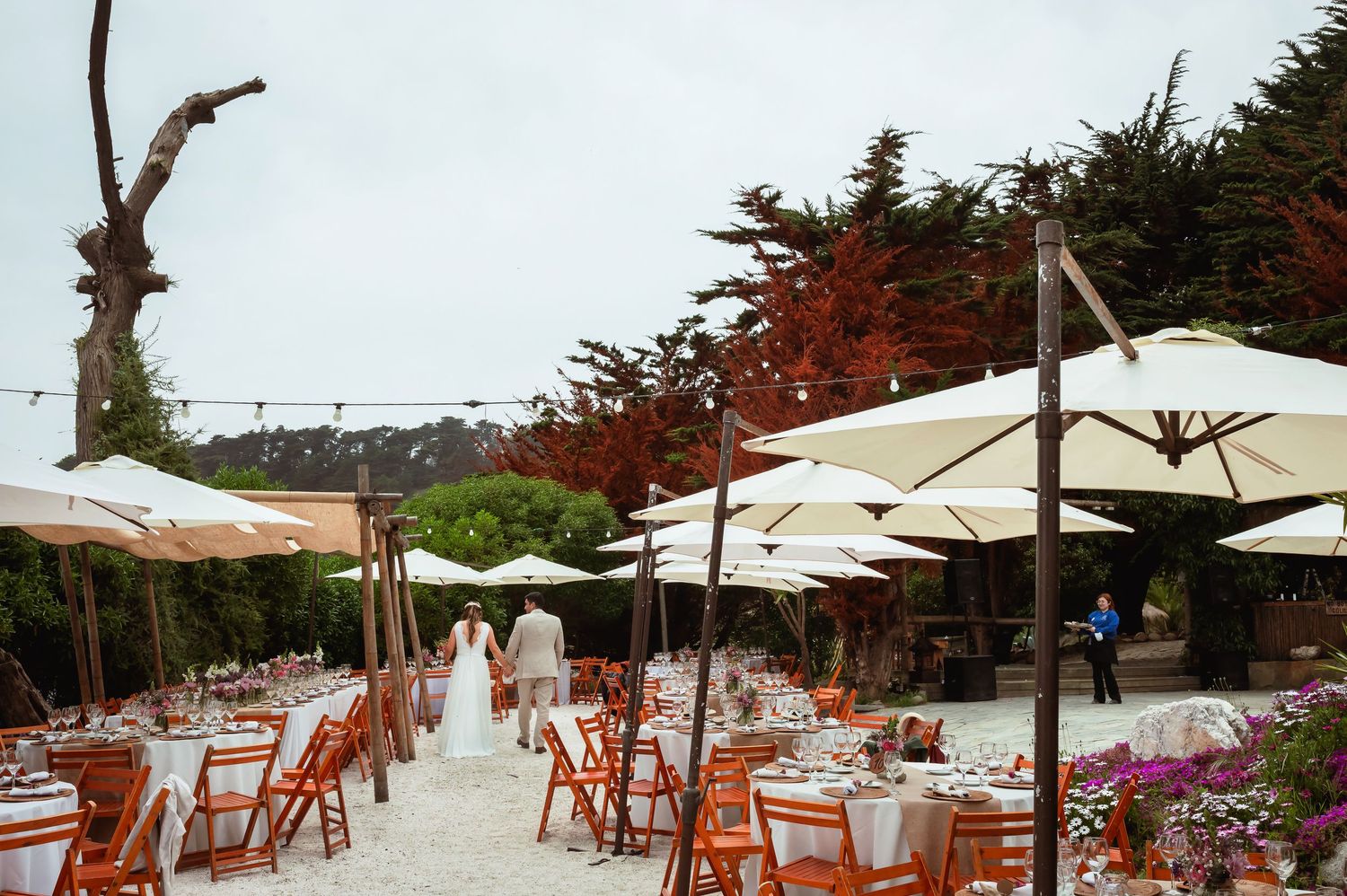 Elegant outdoor wedding reception setup with white umbrellas, orange chairs, and decorated tables in a garden.