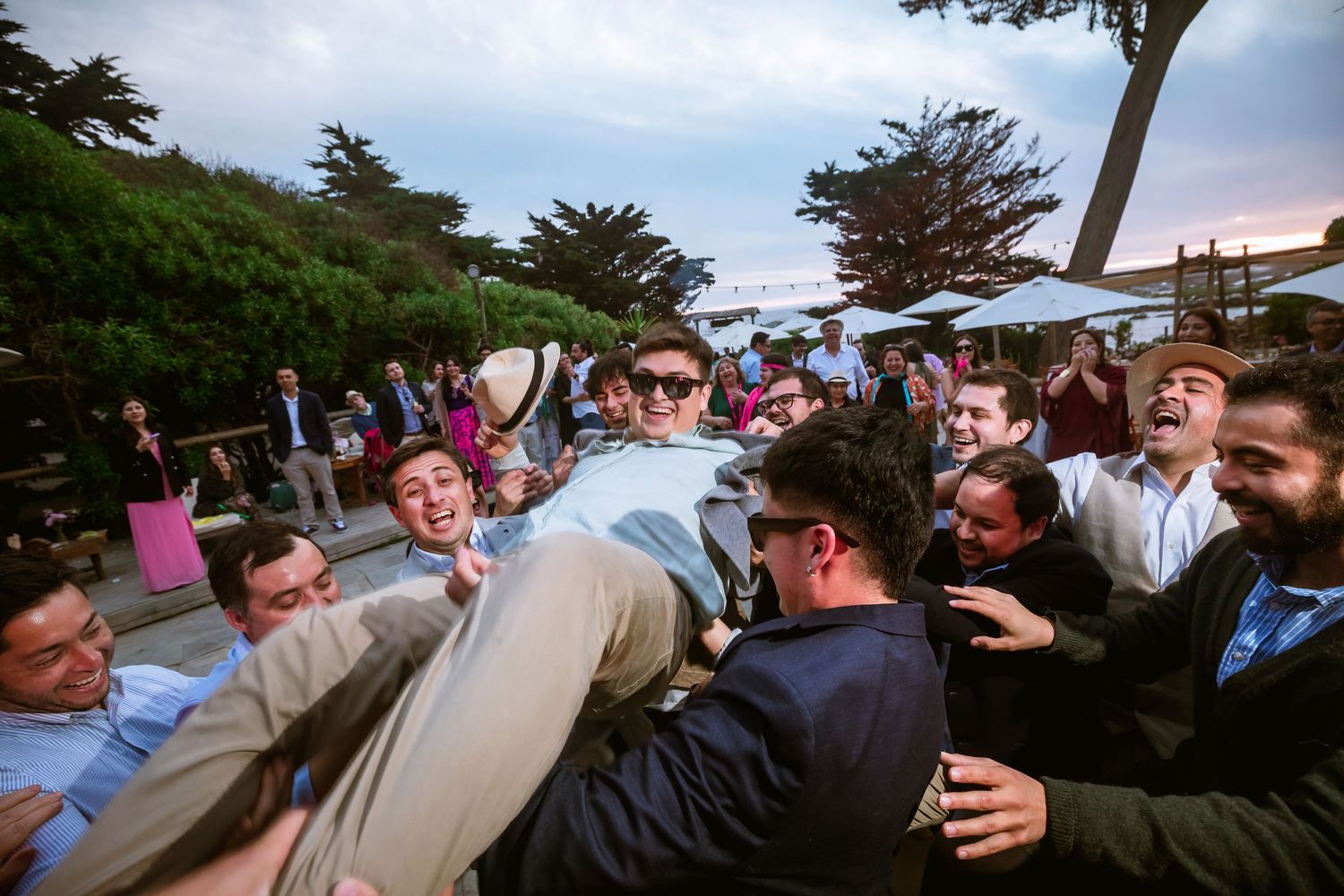 Groom lifted by cheering groomsmen at a lively outdoor wedding reception at dusk.