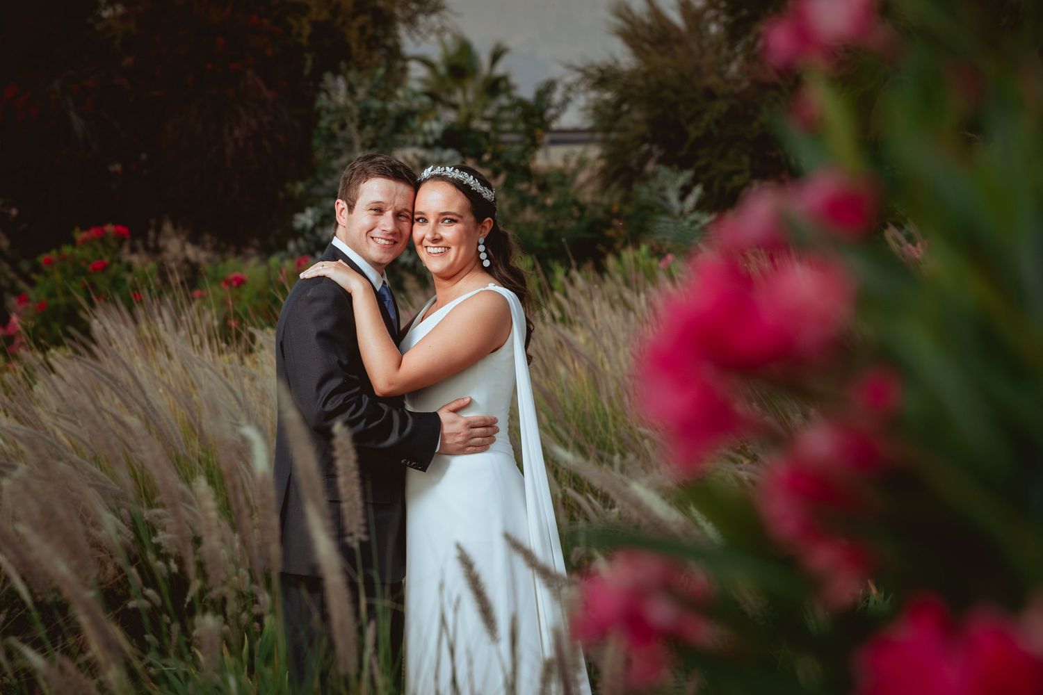 Smiling bride and groom hugging closely surrounded by pink blooms and golden grass.