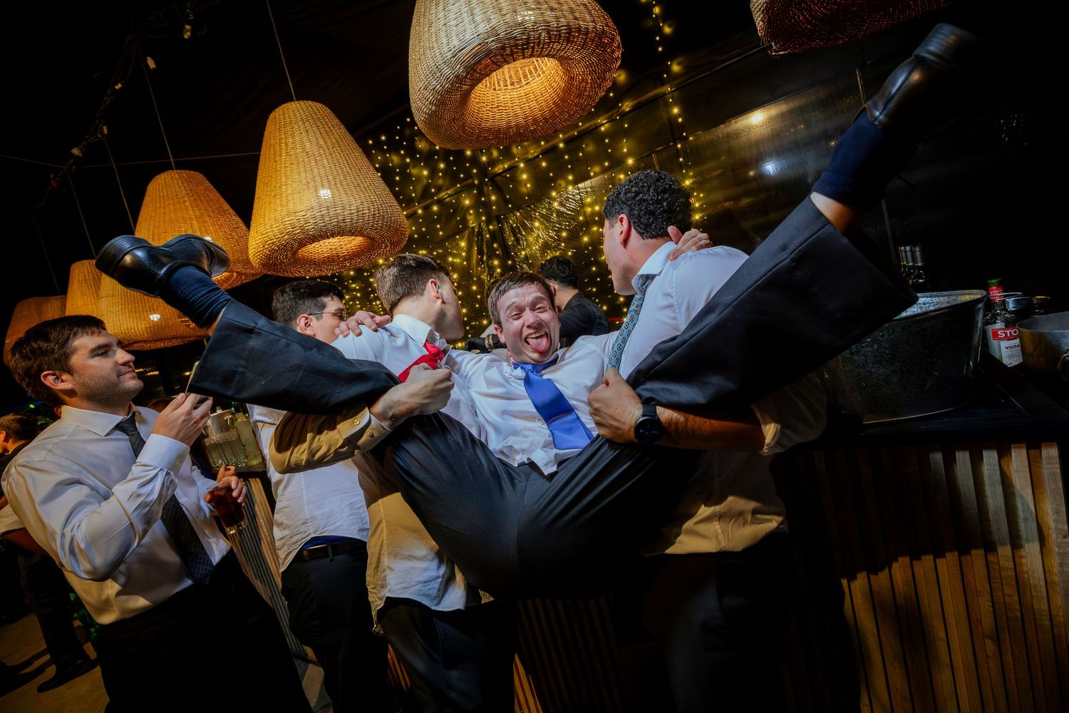 Groomsmen in suits playfully lifting a man in a blue tie at a wedding celebration.