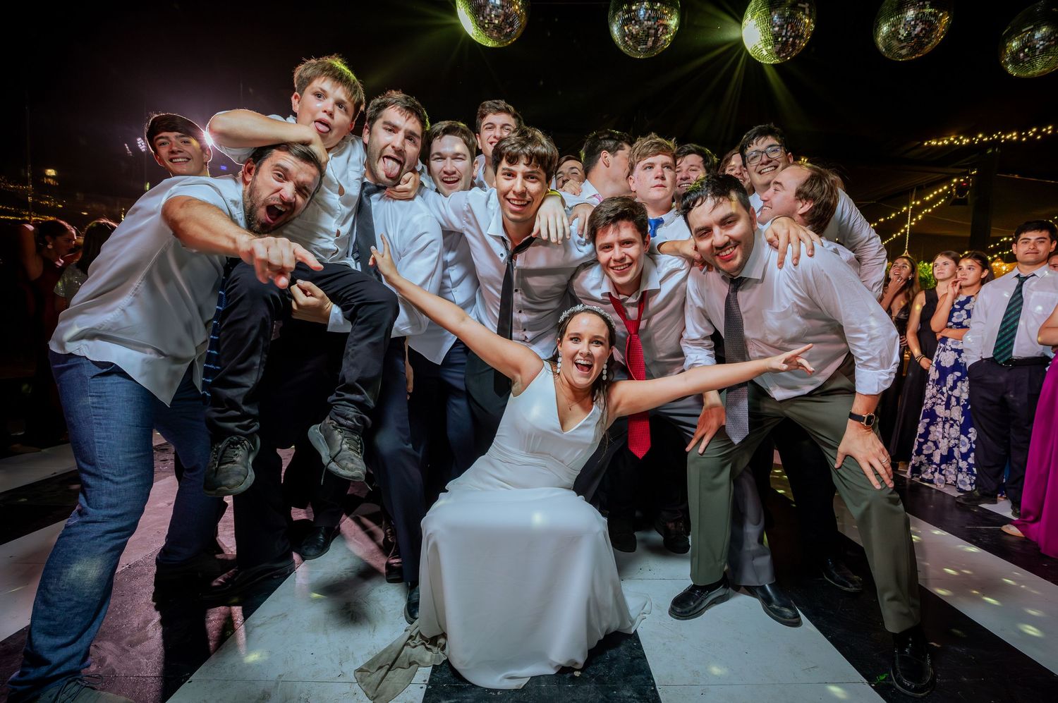Bride in white dress poses with group of young men on wedding dance floor under disco ball.