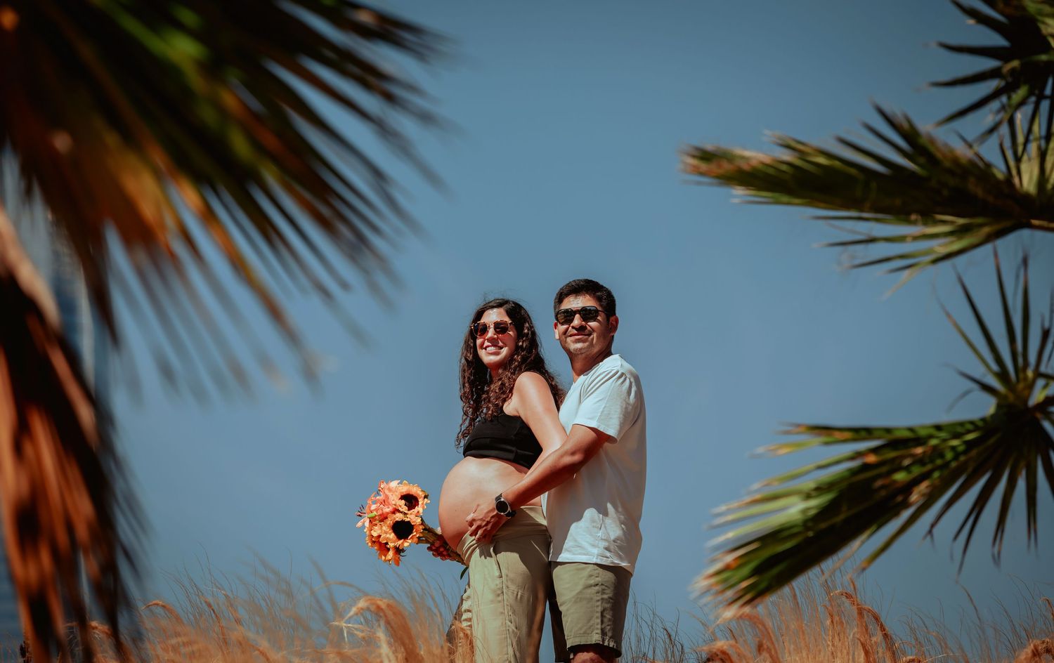 Smiling couple embracing in golden grass field with palm trees, woman holding sunflower bouquet, sunny day.