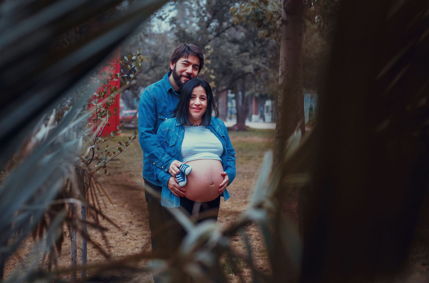 Smiling pregnant couple posing among foliage in a park, woman holding baby shoes on belly.