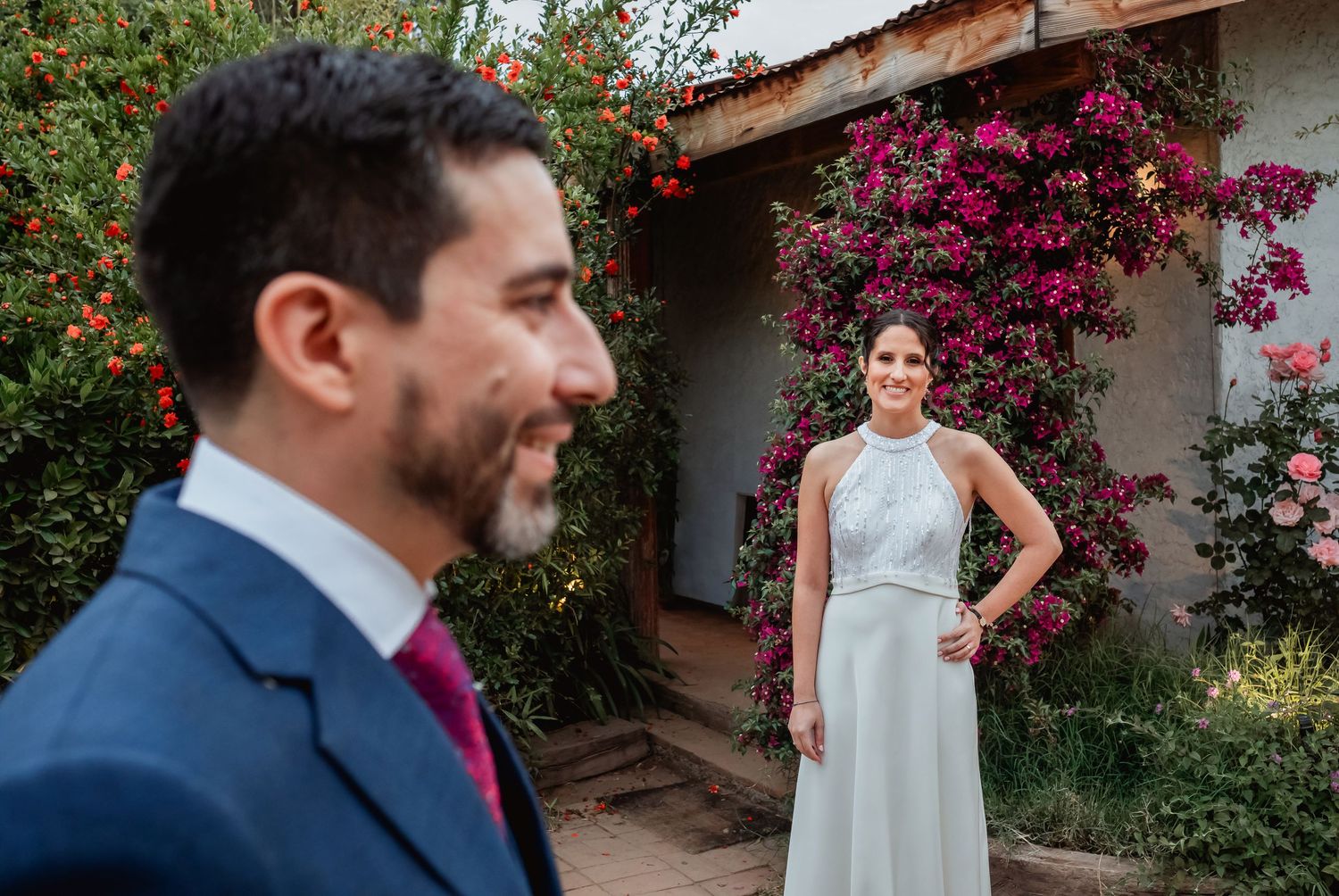 Groom smiles as bride in halter wedding gown stands near vibrant bougainvillea wall.