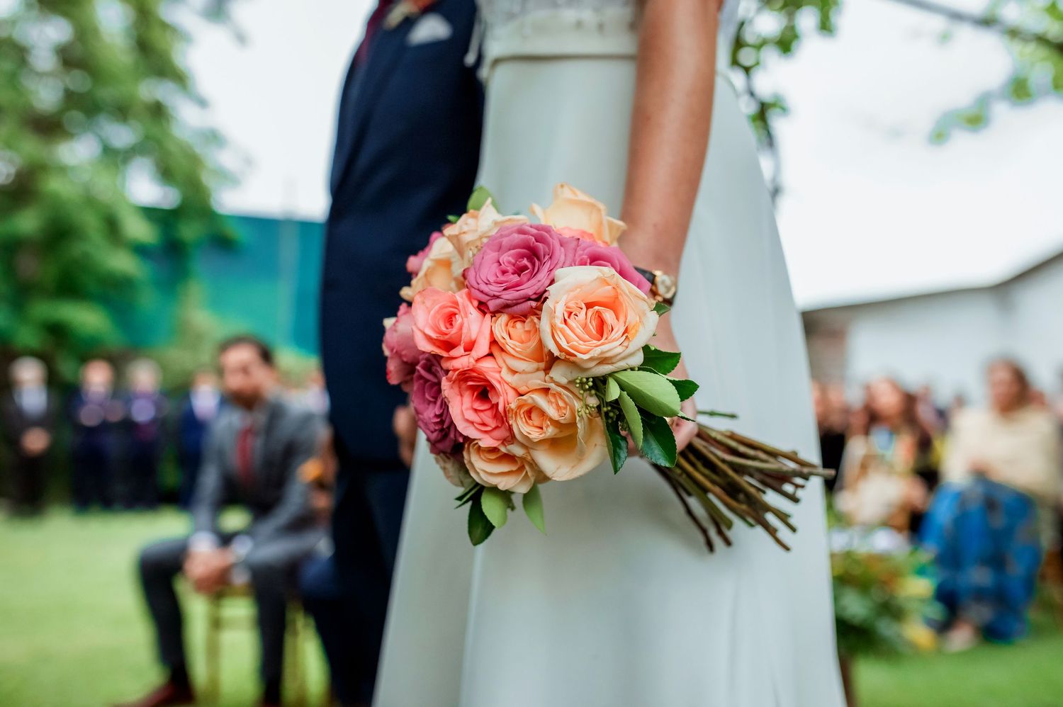 Bride holds colorful pink and orange rose bouquet during outdoor wedding ceremony.