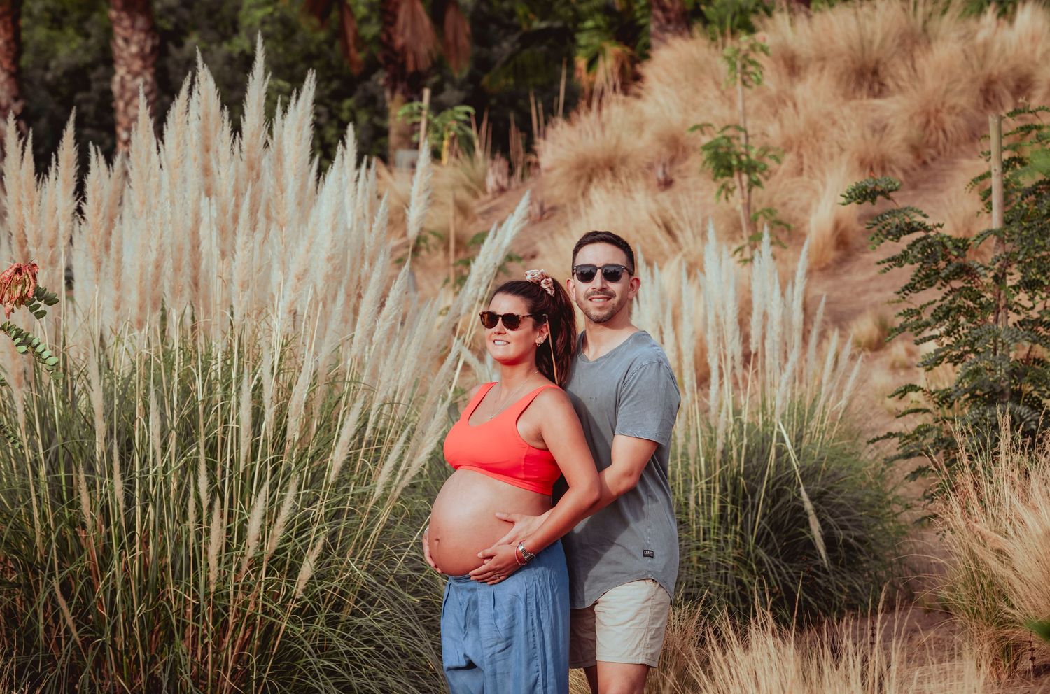 Expectant couple standing close together near tall pampas grass and palm trees in warm sunlight.