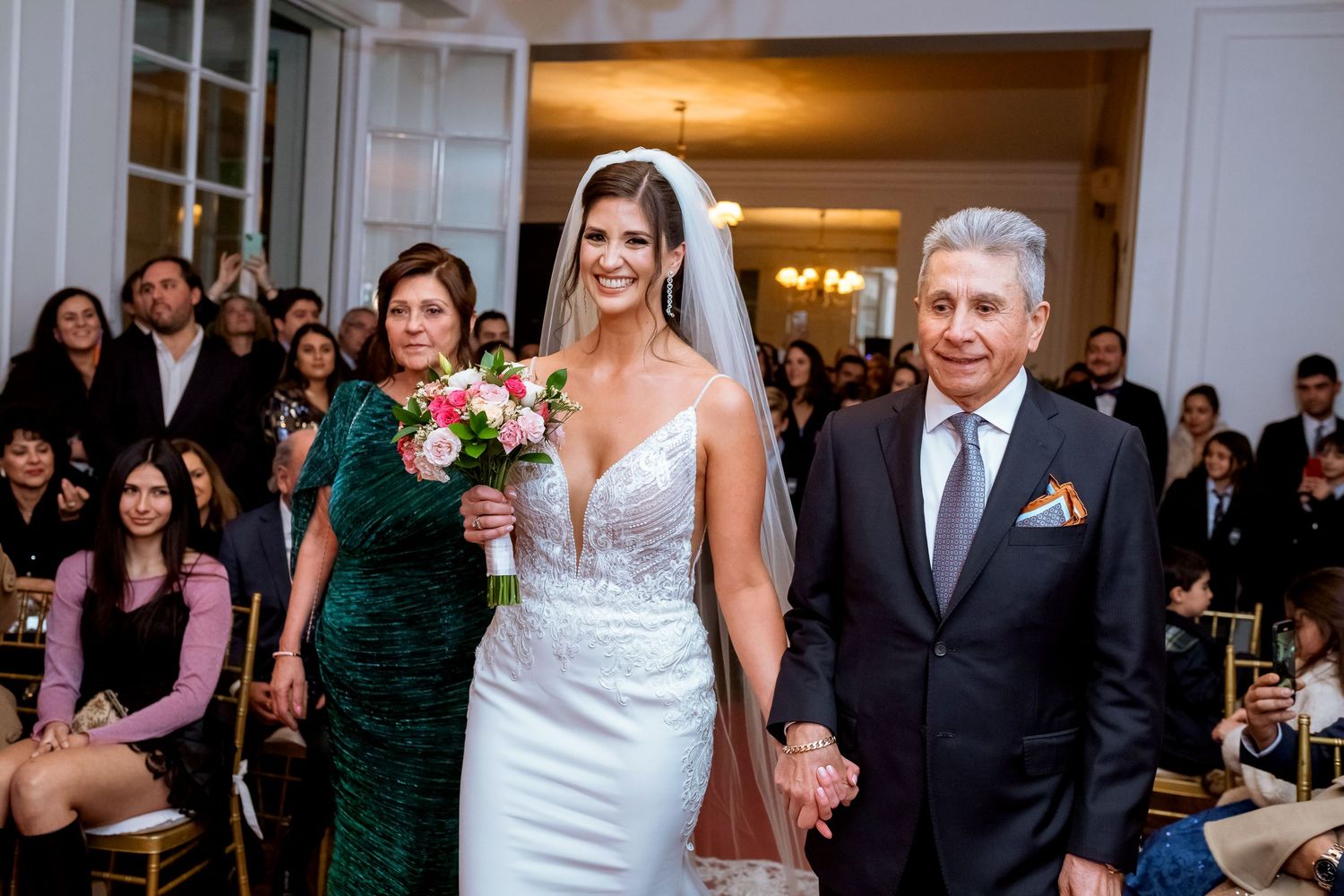 Bride in white wedding gown walks down aisle with parents during elegant indoor wedding ceremony.