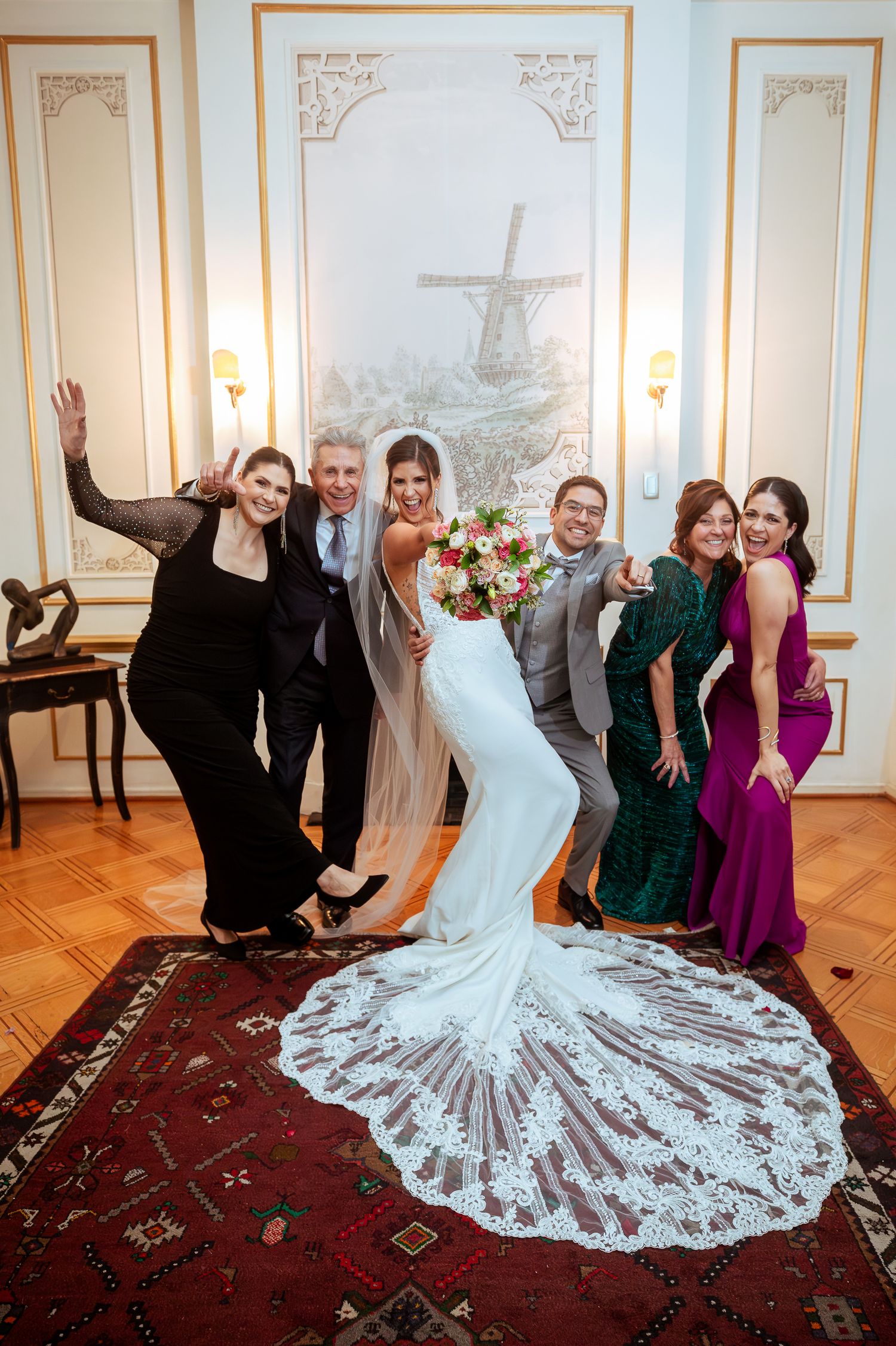 Bride in lace gown with bouquet poses with wedding party in elegant room featuring windmill mural.