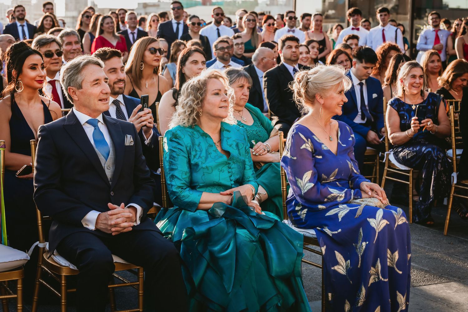 Wedding guests seated in rows, including woman in teal gown, at outdoor ceremony.