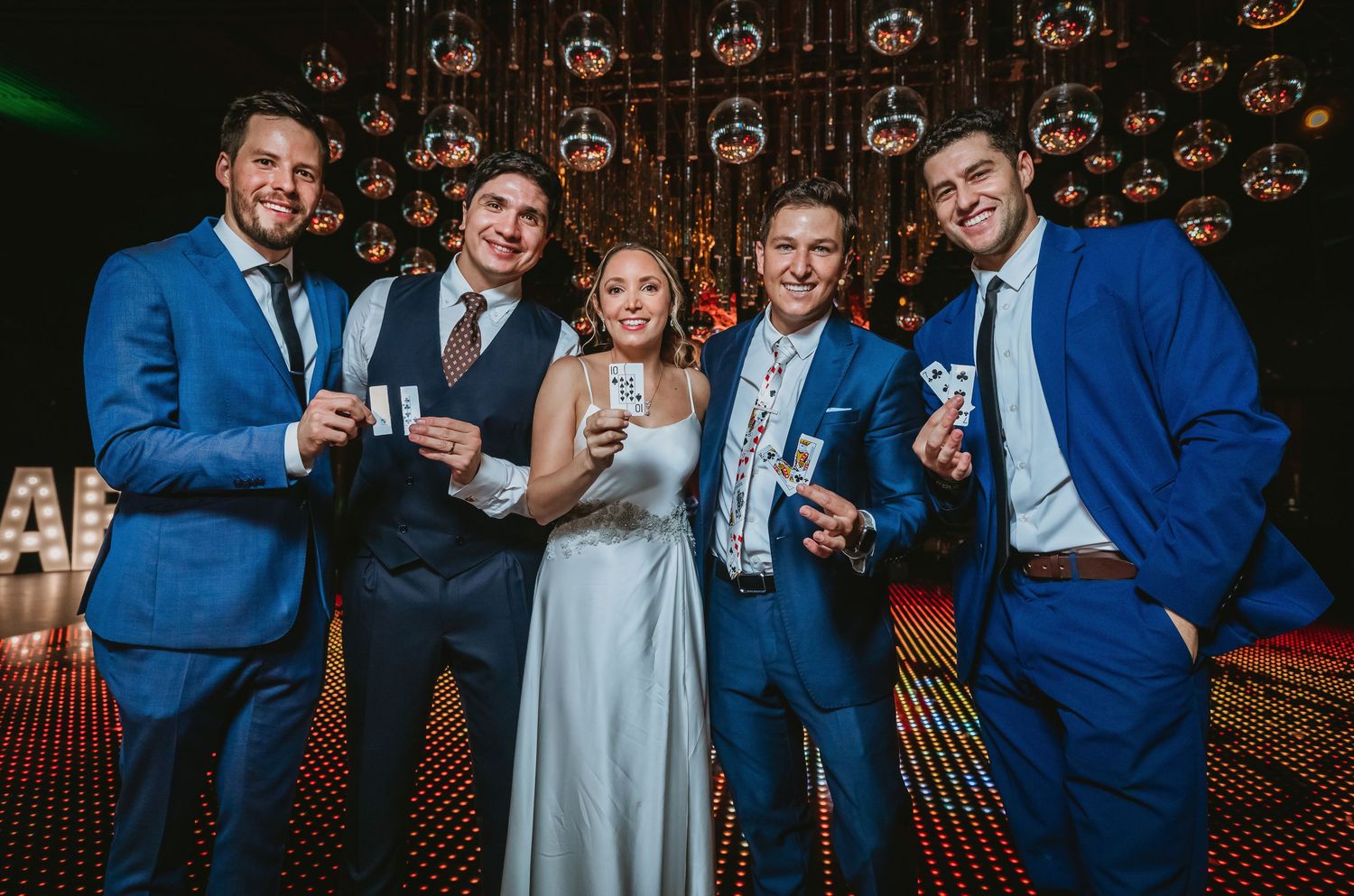 Five wedding guests in blue suits and bridal gown pose together holding drinks at a festive reception venue.