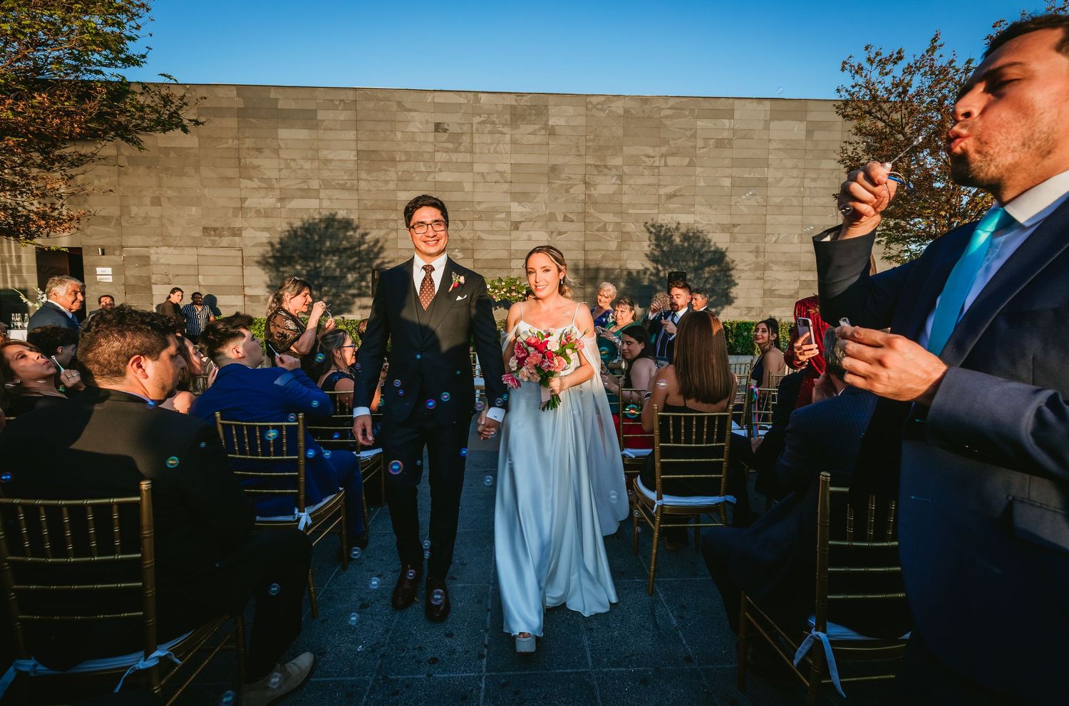 Couple walks down aisle as guests toss confetti at outdoor wedding ceremony.