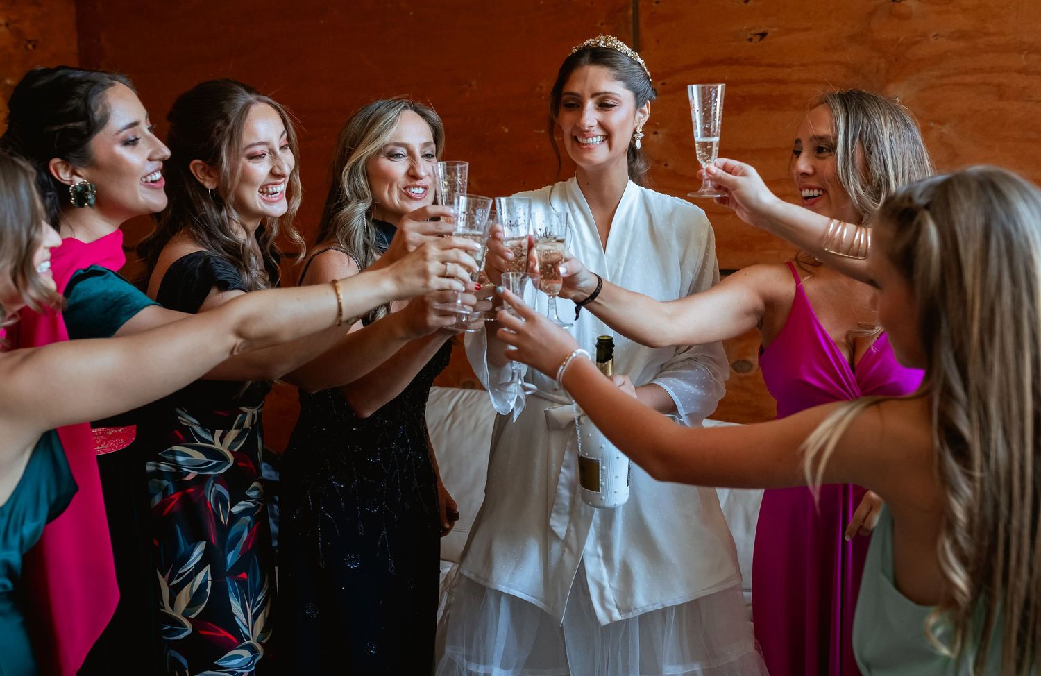 Bride in white robe toasting champagne with bridesmaids in colorful dresses during wedding day celebrations.